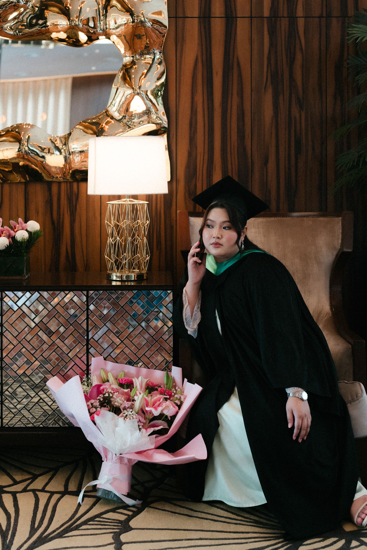A graduate in academic regalia poses with pink flowers against wood paneling and decorative lighting.