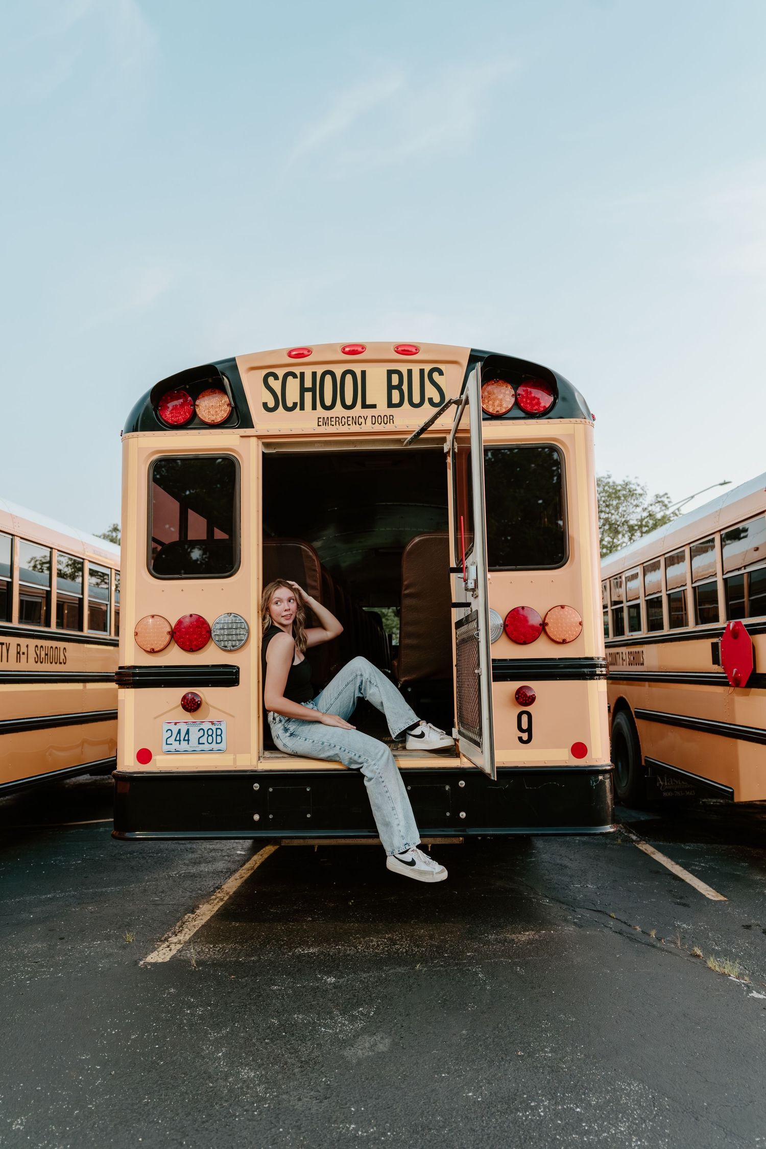 A student poses casually in the doorway of a classic yellow school bus in a parking lot.