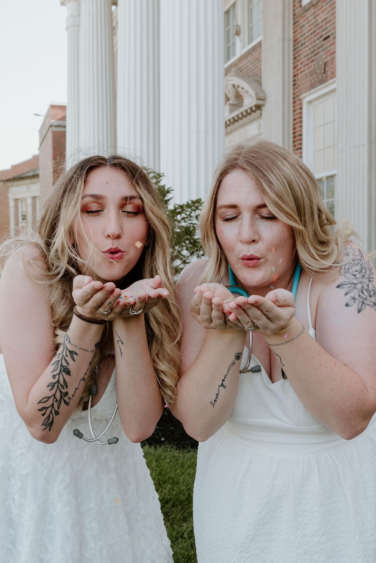 Two women in white dresses making blowing gestures with cupped hands outdoors by a columned building.