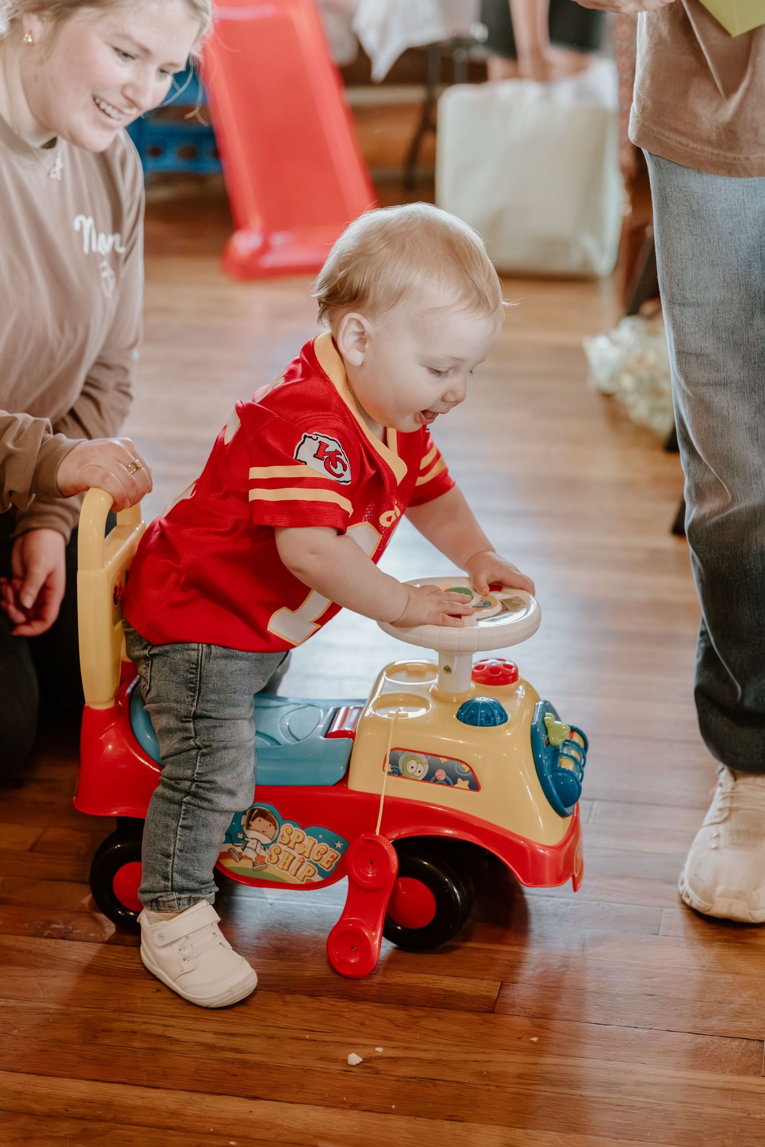Small child in red shirt plays with a vintage-style ride-on toy car on hardwood floors.