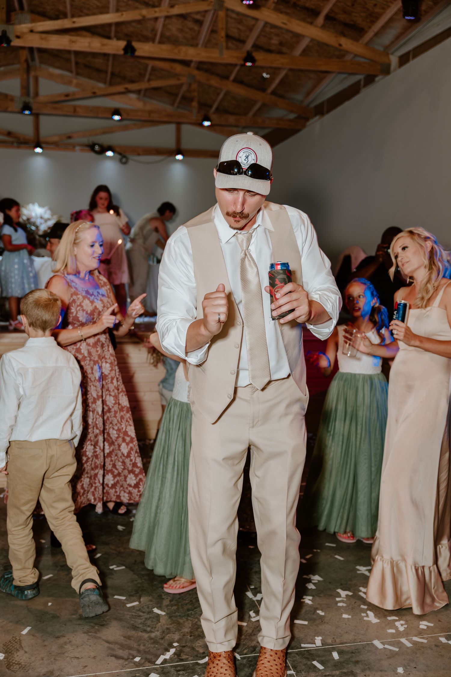 Groomsman in tan vest and backwards cap dances energetically holding a drink at lively indoor wedding reception.