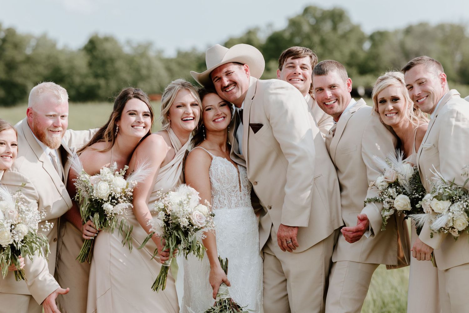 Wedding party in champagne attire poses outdoors, bride in white lace gown holding bouquet, groom in tan suit and cowboy hat.