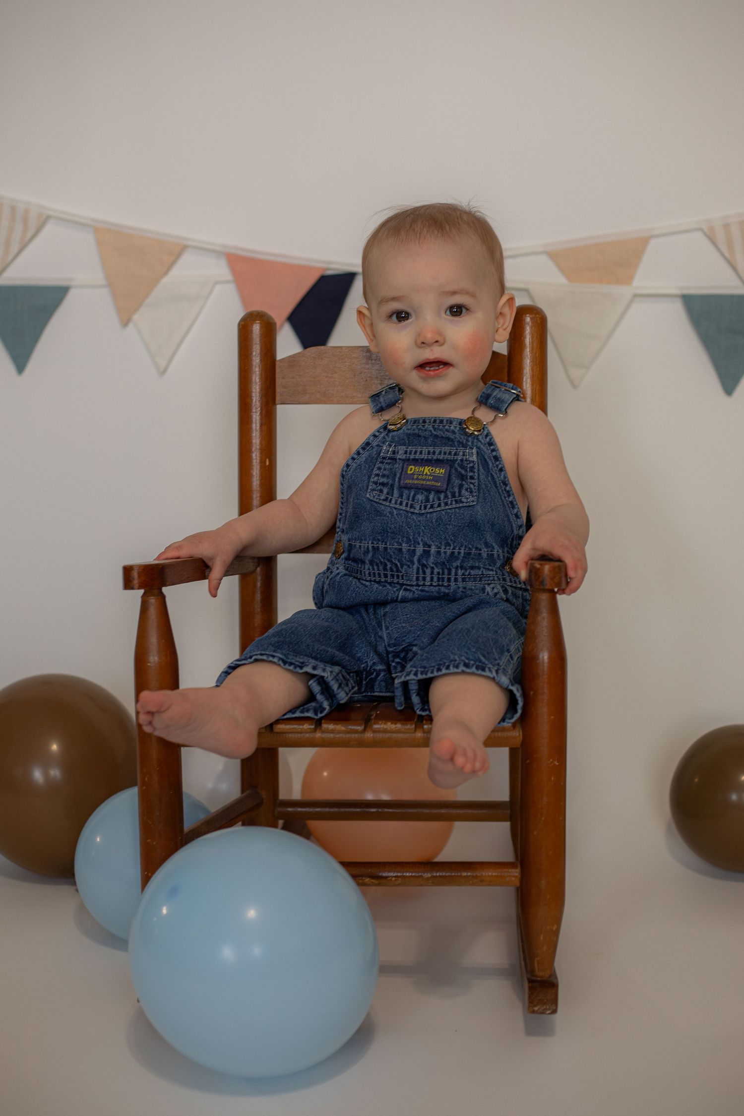 Baby in denim overalls sits in wooden rocking chair with pastel balloons and bunting decorations.