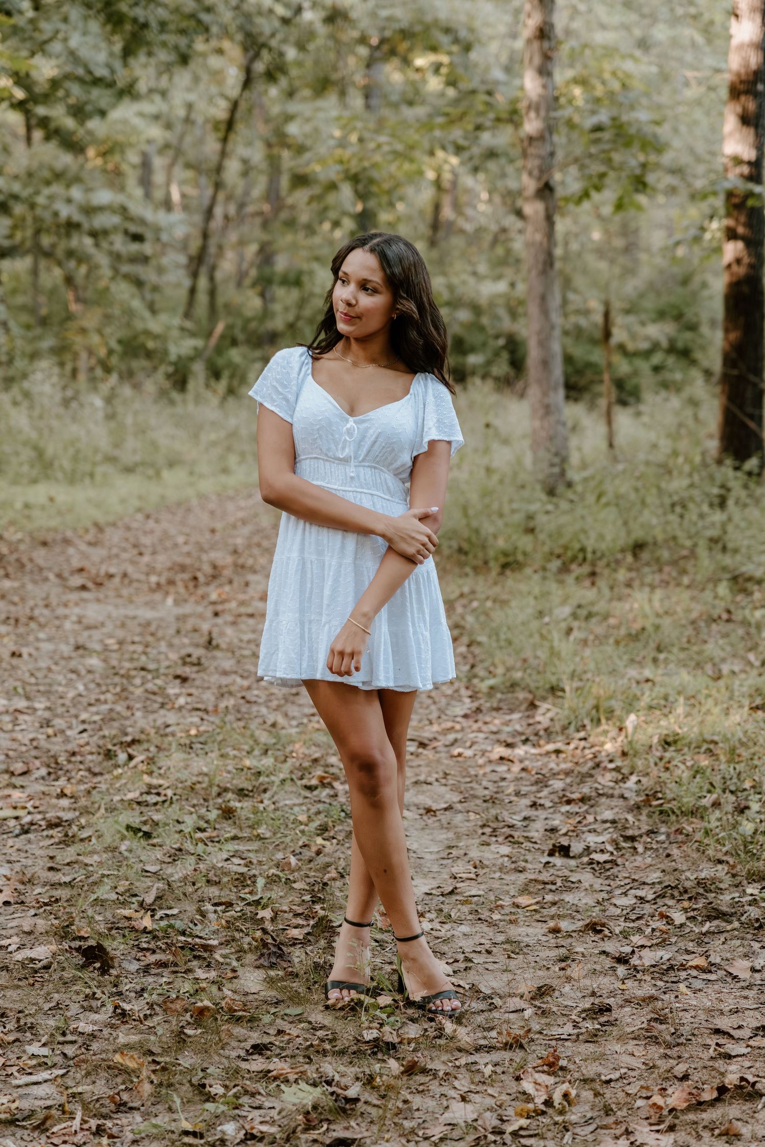 A figure in a white summer dress poses playfully in a wooded area covered with fallen leaves.