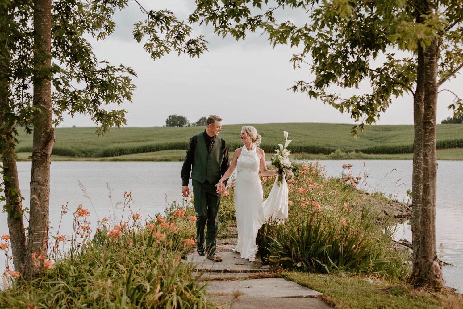 Bride and groom walking hand-in-hand along a scenic lakeside garden path surrounded by lush greenery.
