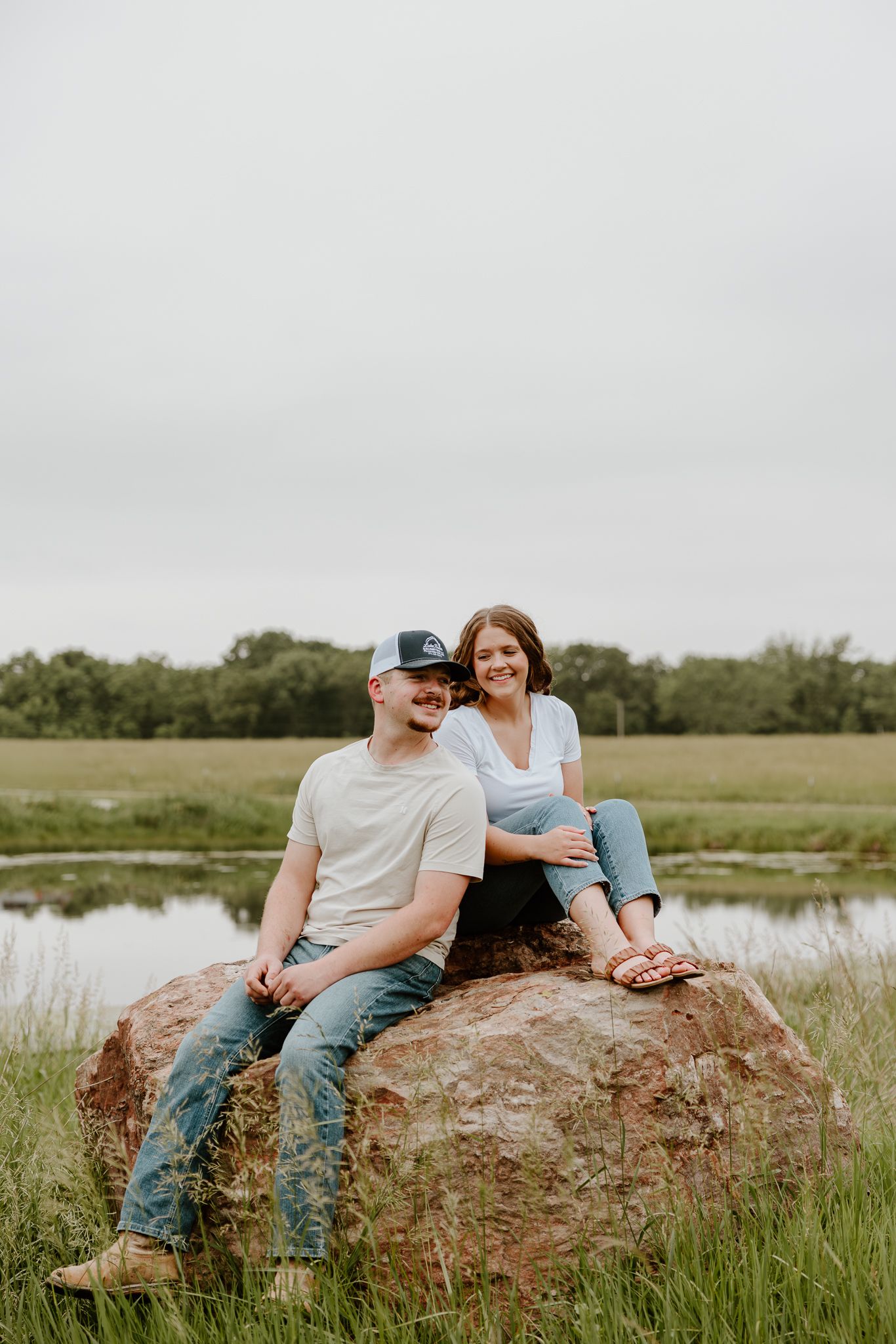 Couple sitting together on a large rock in a green field with a pond in the background.