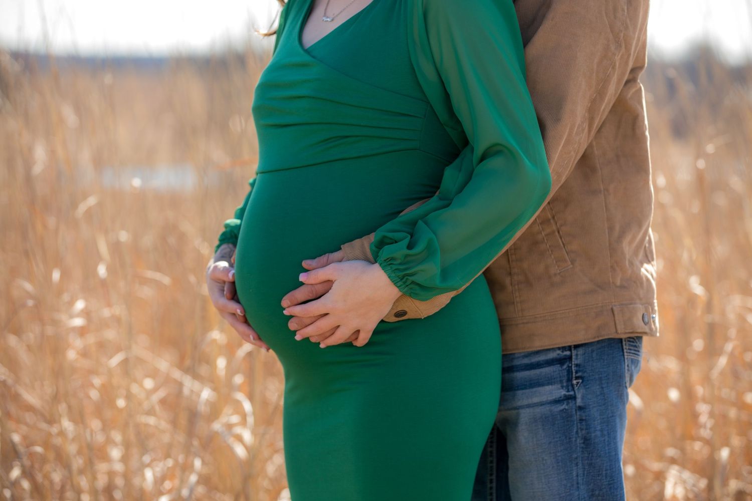 Two people embrace while showing off a baby bump in a flowing green dress against tall grass.