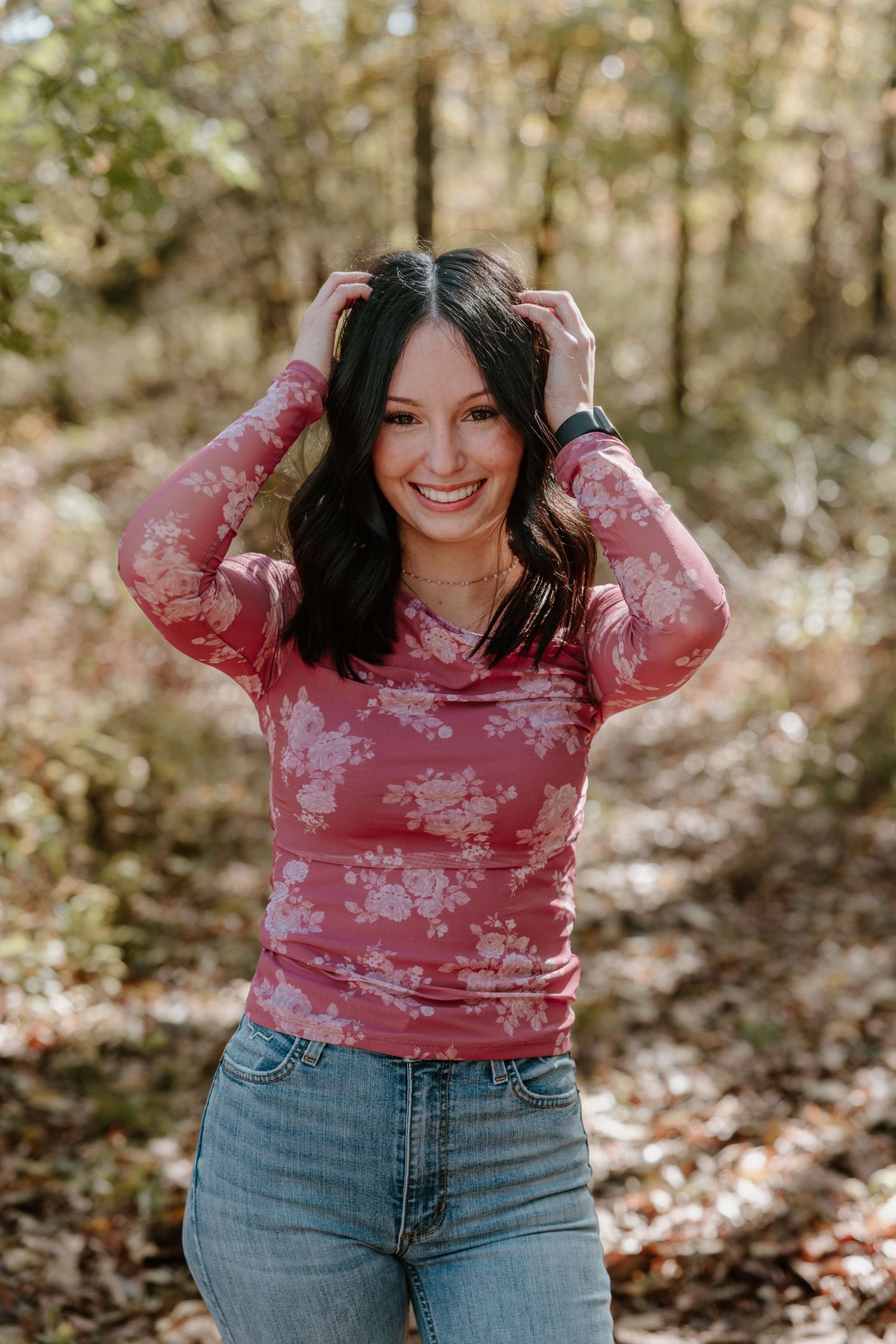A woman wearing a pink floral sweater and jeans smiles while posing in a wooded area with fallen leaves.