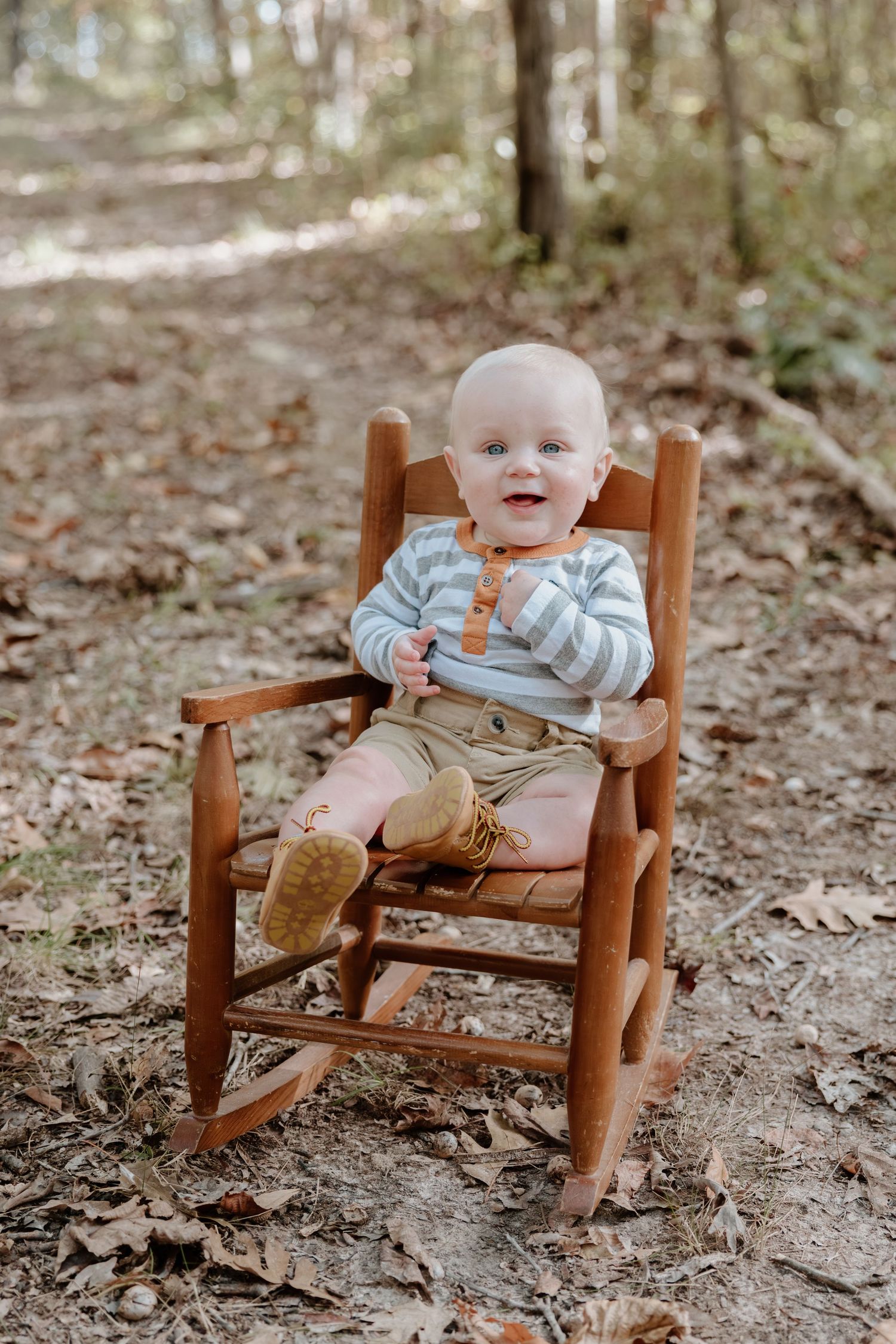 A small child sits in a wooden rocking chair on a forest floor covered with fallen leaves.