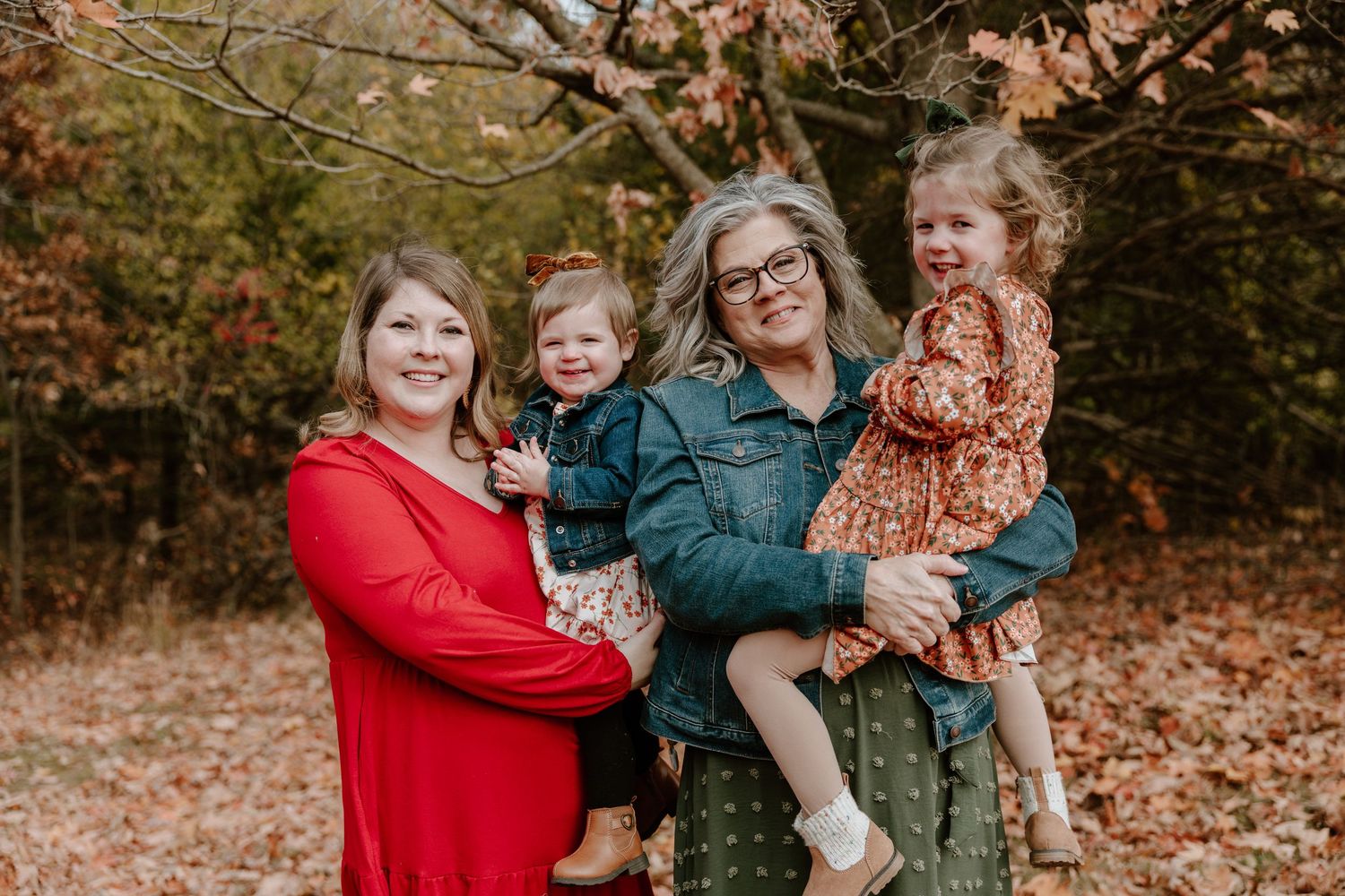 A family wearing red and green sweaters poses together in an autumn forest setting with fallen leaves on the ground.