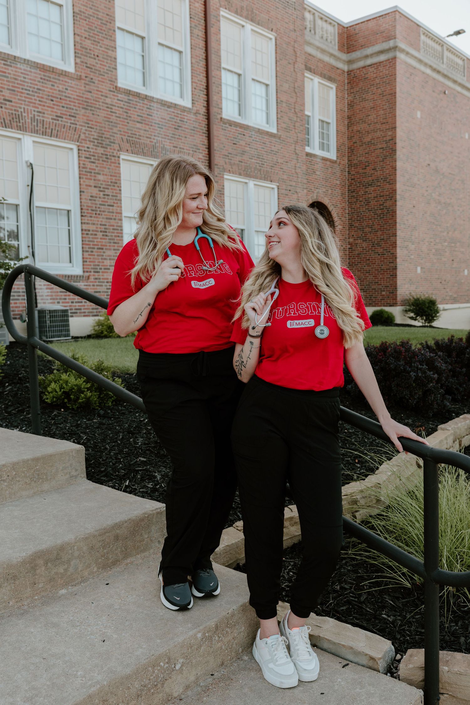 Two friends wearing matching red shirts and black pants laugh together while standing on brick building steps outdoors.