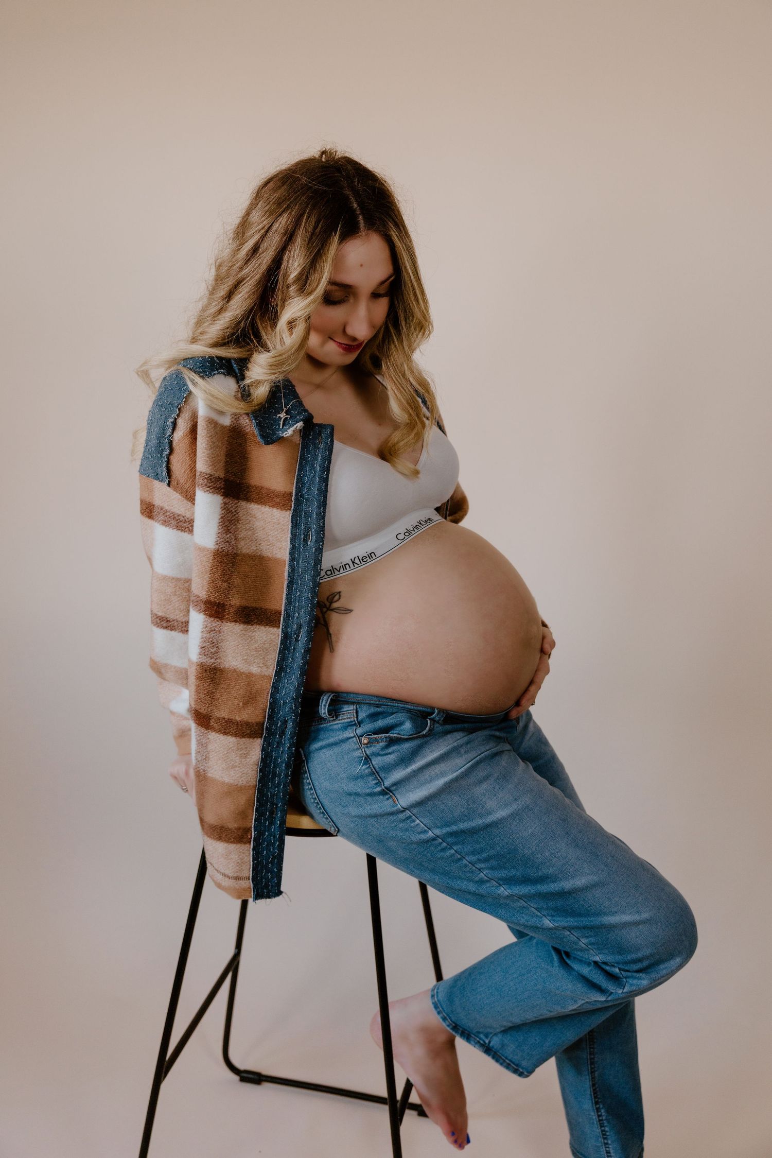 A pregnant woman in casual denim and a striped cardigan sits on a stool against a neutral backdrop in a studio setting.
