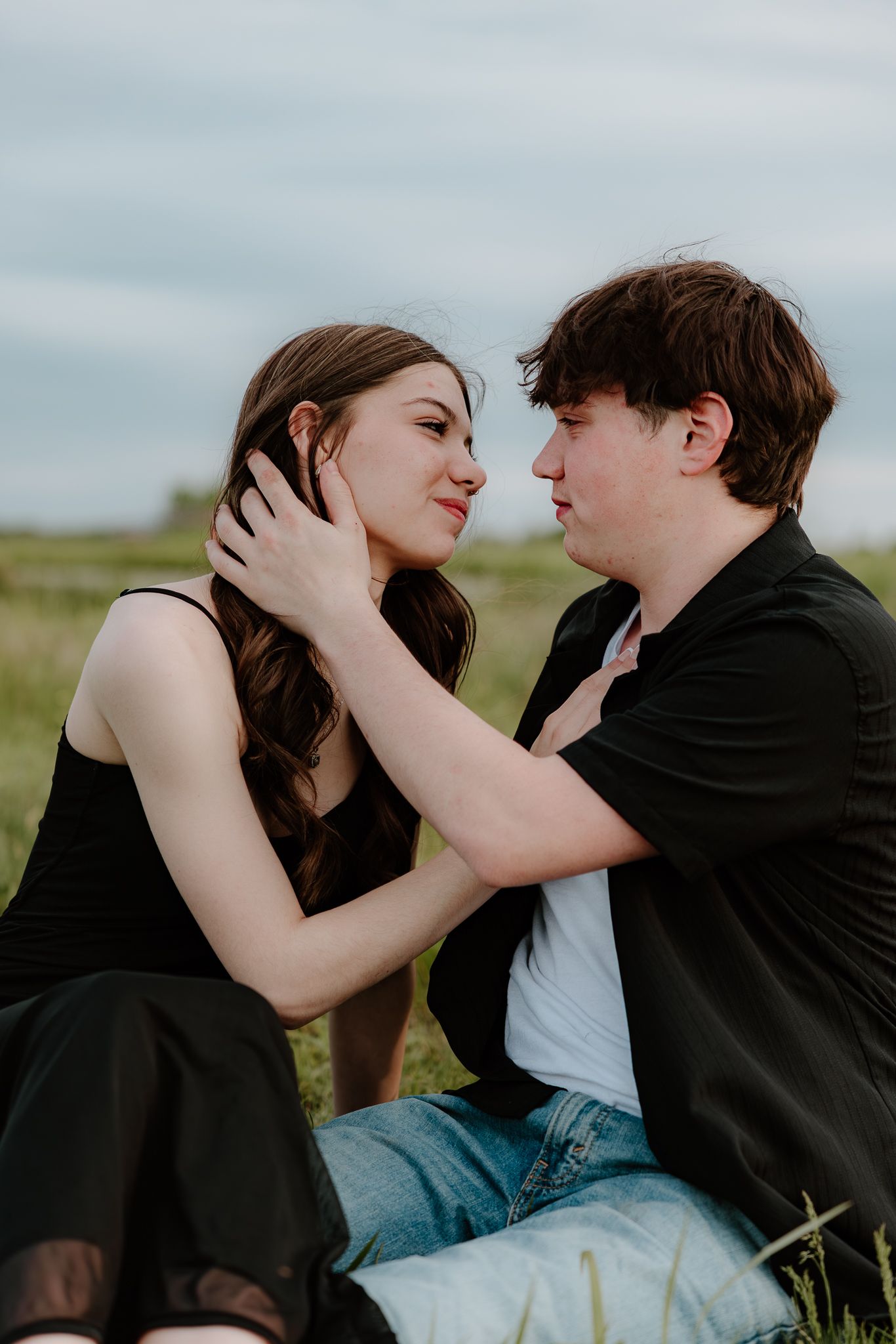 A young couple sits in a grassy field, gazing into each other's eyes in a tender romantic moment.