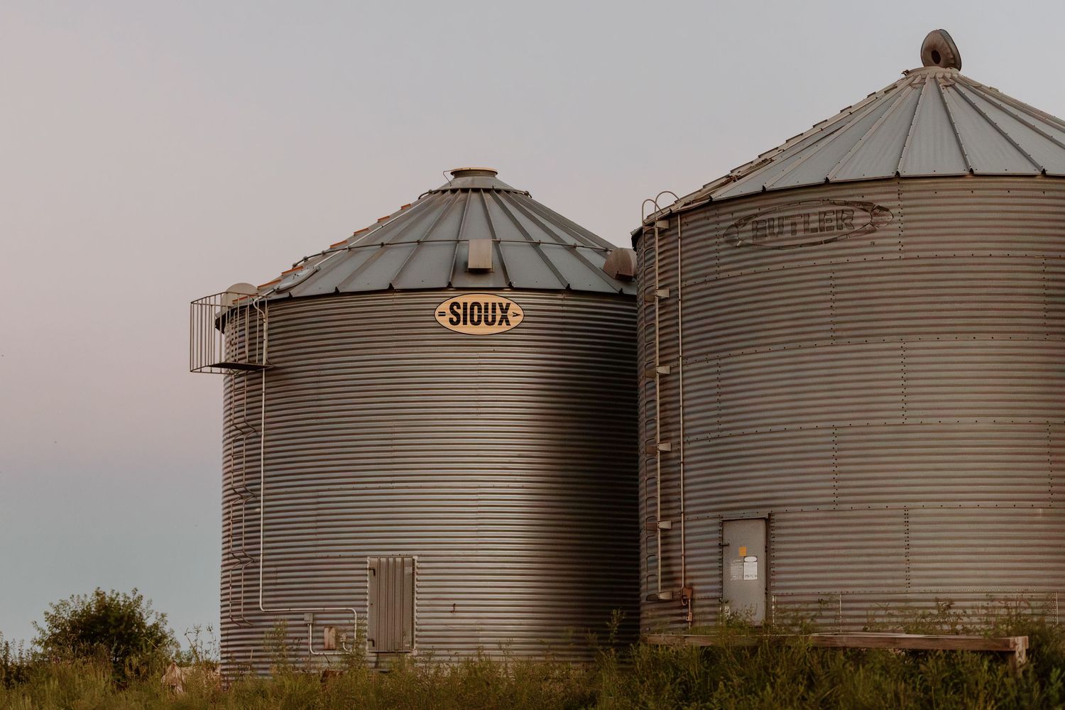 Two large Sioux brand steel grain storage silos on a farm at dusk with green grass below.