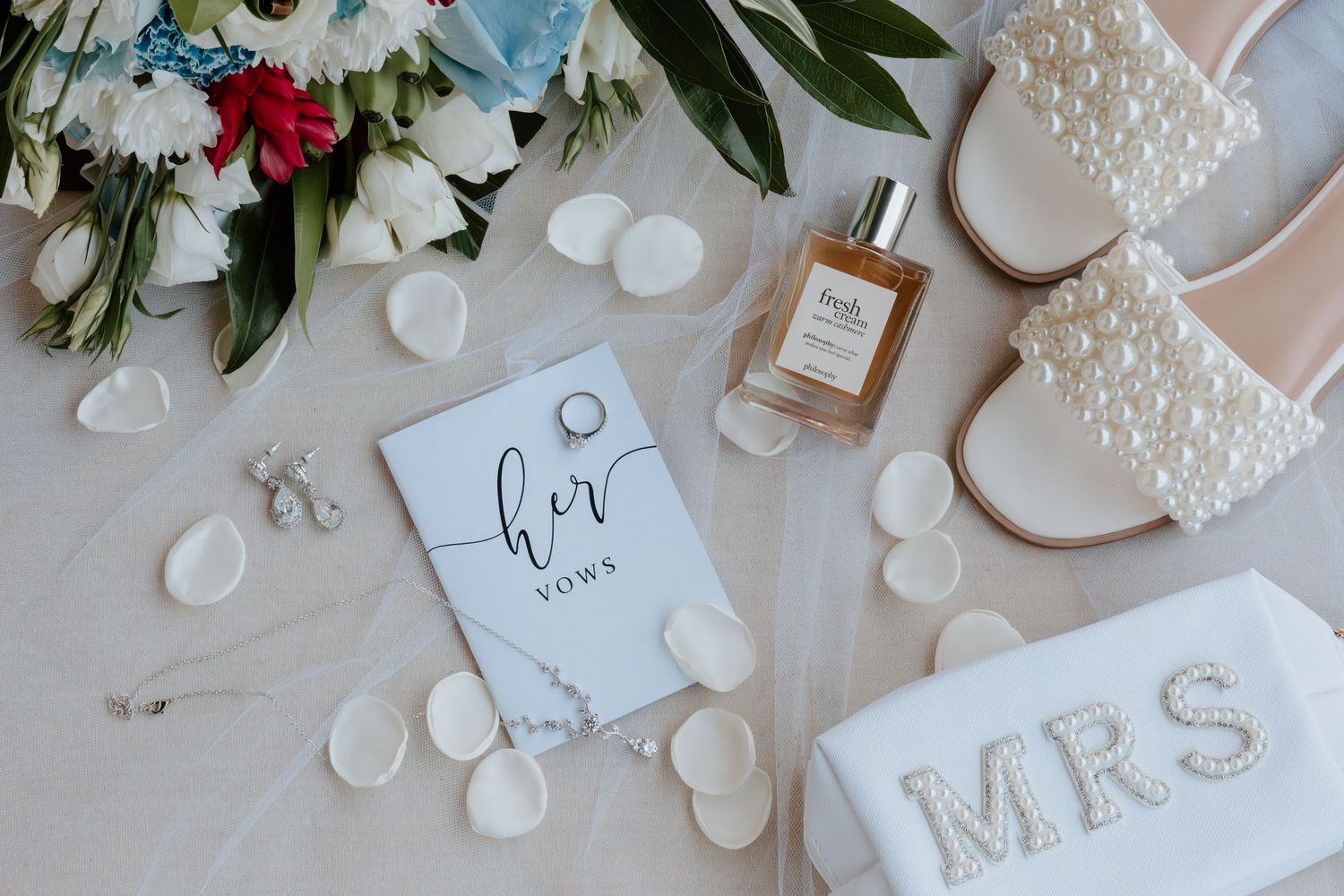 Flat lay of bridal accessories including vow book, pearl heels, perfume, and MRS clutch.