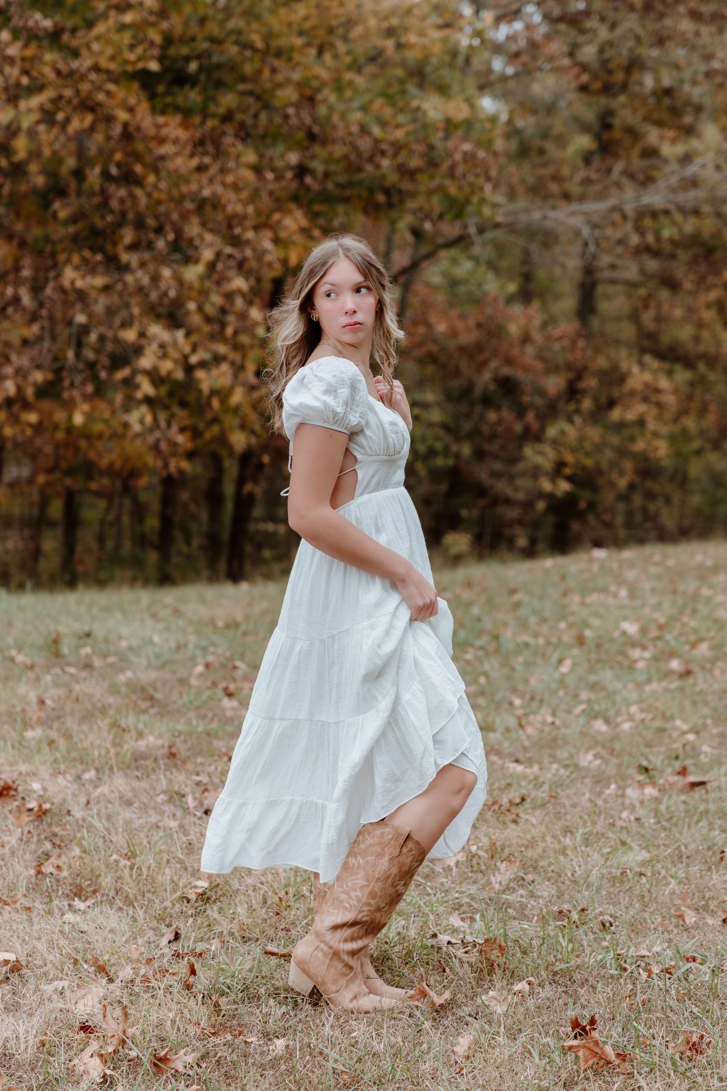 A child in a white dress and brown boots twirls playfully in a fall setting with autumn leaves on the ground.