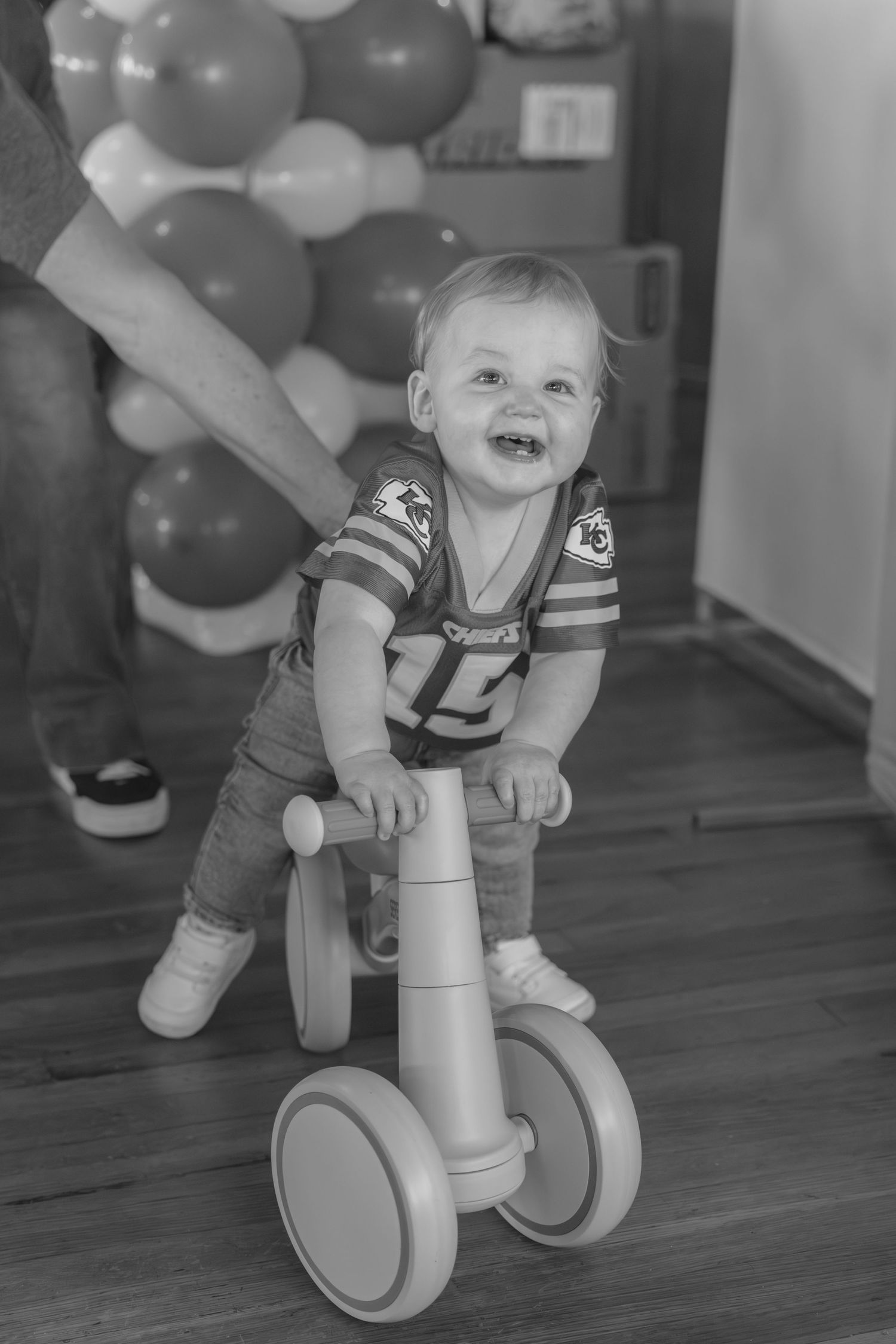 A happy baby rides on a minimalist white balance bike against a hardwood floor with balloons in the background.