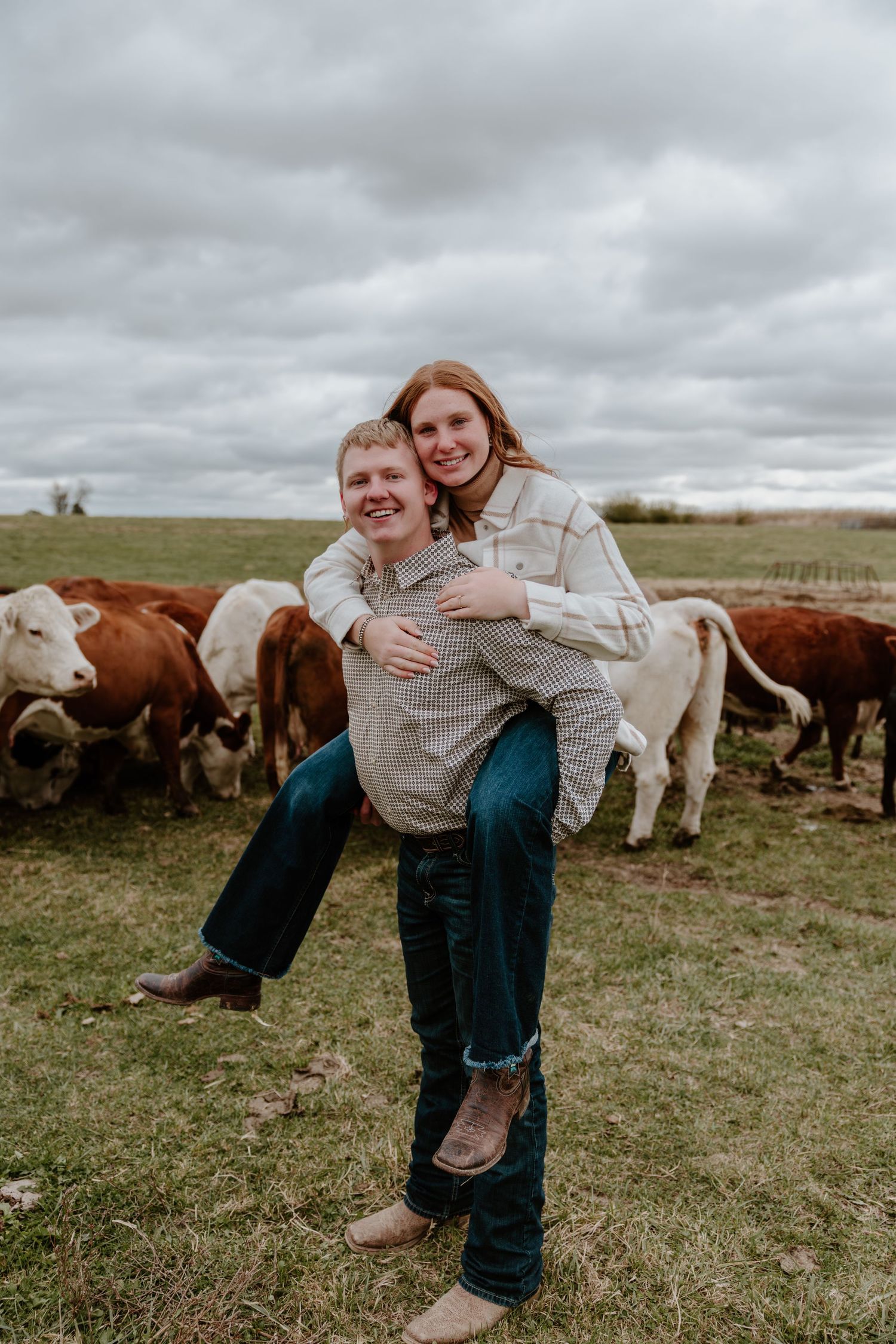Playful couple posing in a field with grazing cattle and cloudy skies in the background.