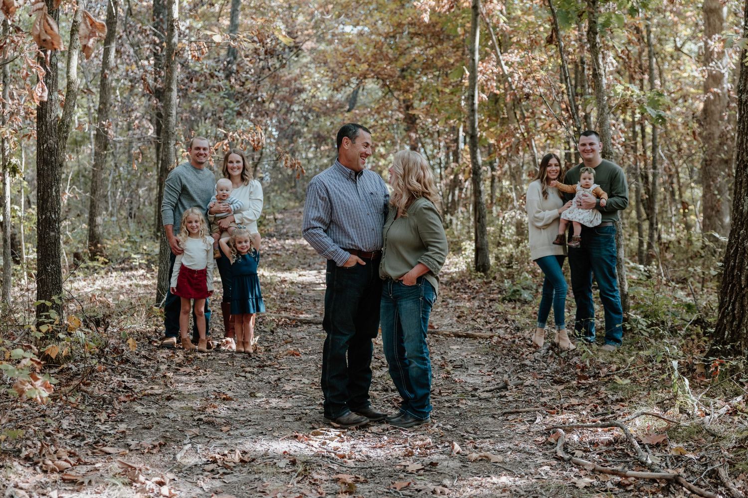 People gather in a wooded area during autumn with fallen leaves scattered on the ground.