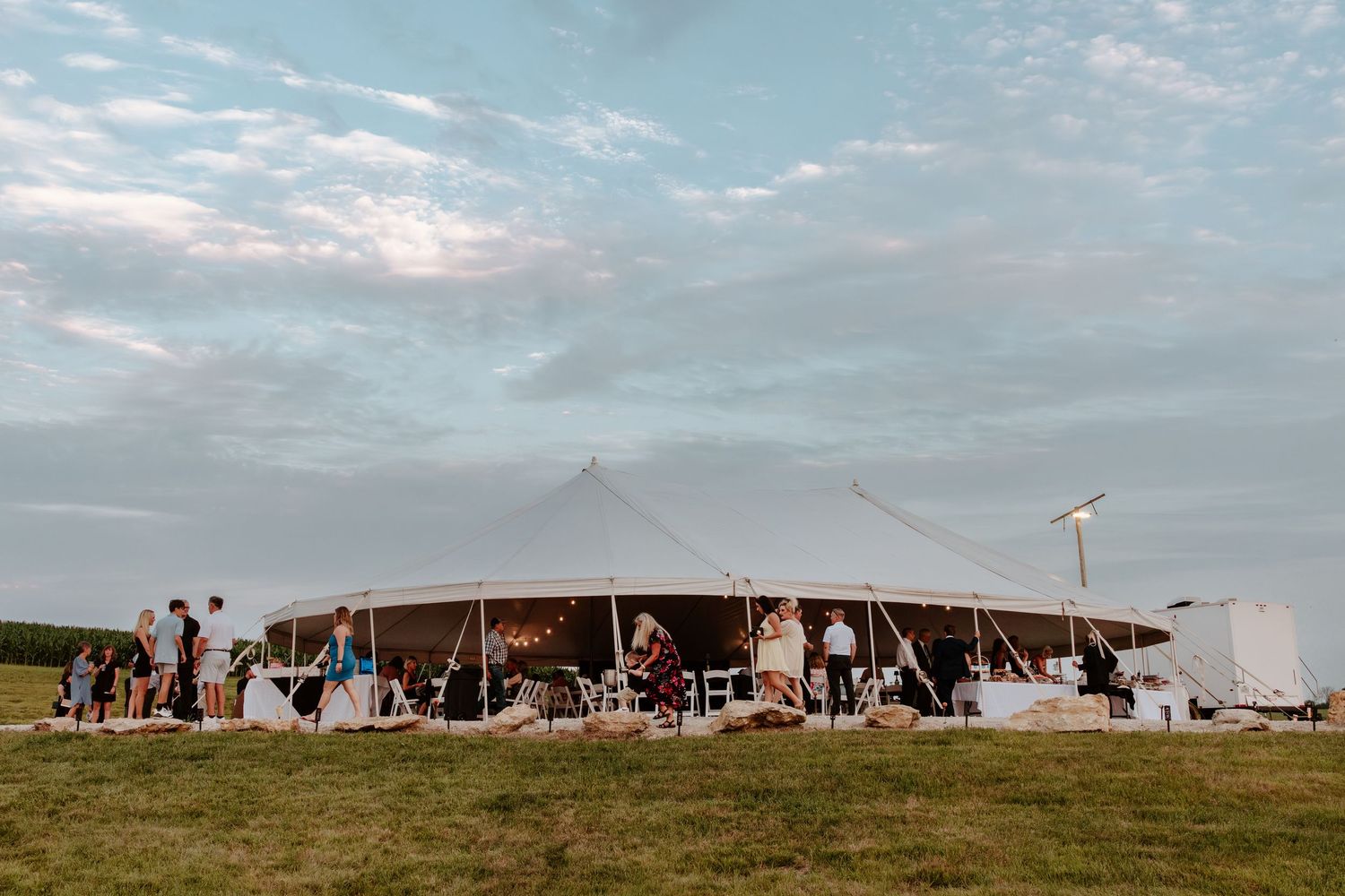 Large white sailcloth wedding tent on green field at dusk with guests mingling outside.