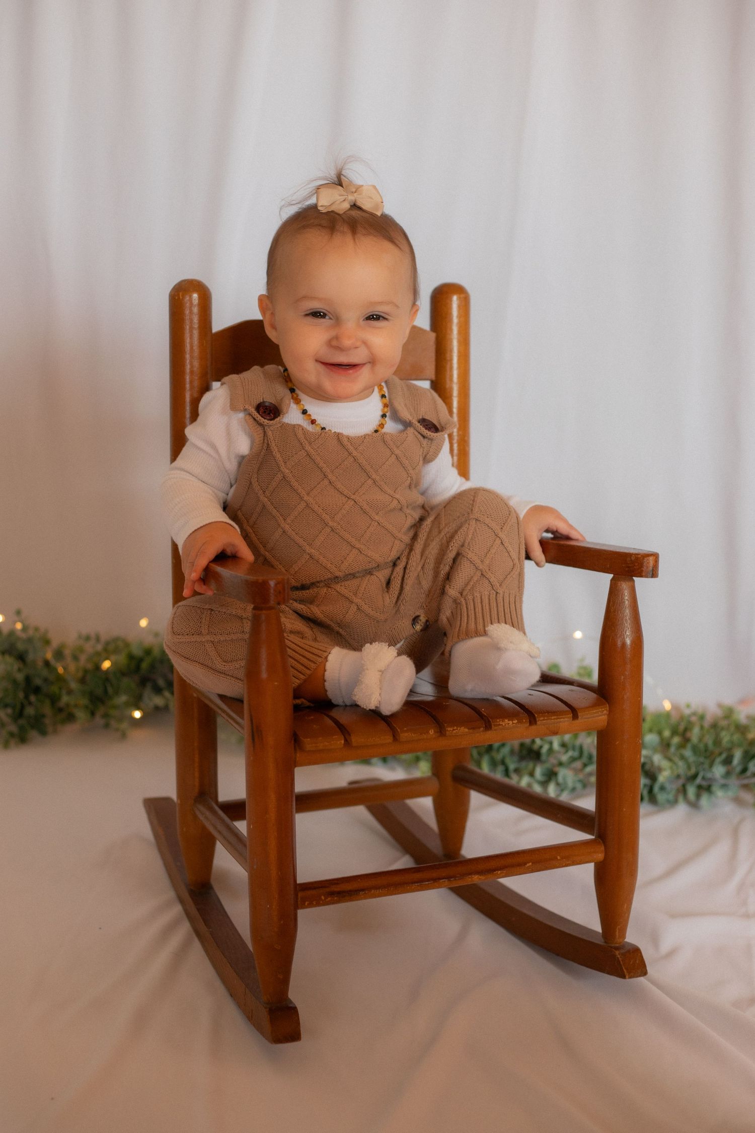 Baby wearing knit outfit sitting in wooden rocking chair with holiday lights behind.