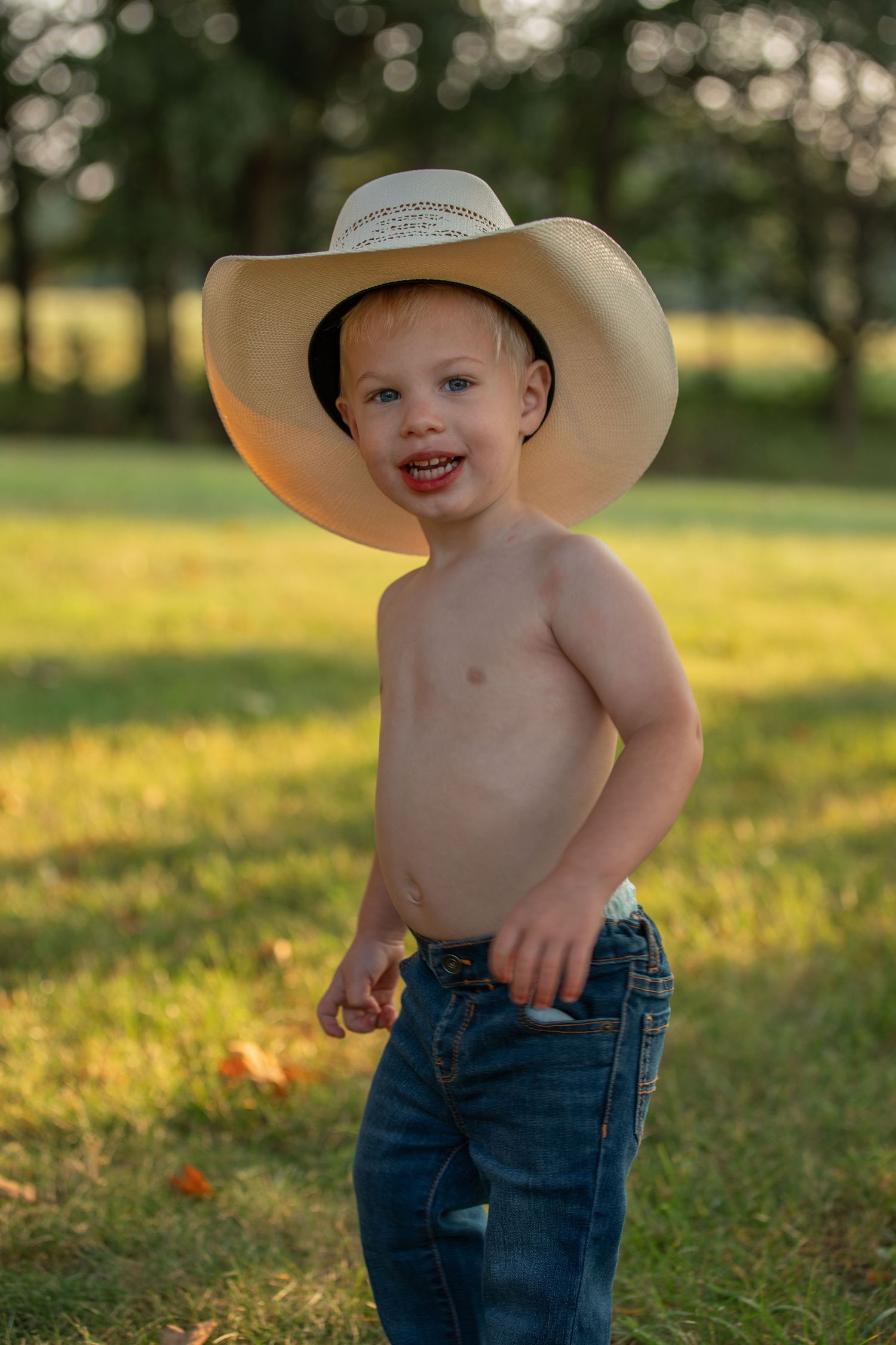 Smiling child wearing cowboy hat and jeans poses in sunny backyard.