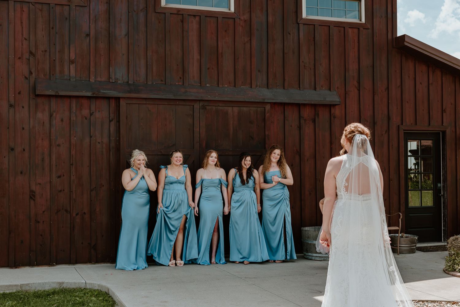Bride in white gown and veil stands before cheering bridesmaids in blue dresses outside a rustic red barn.