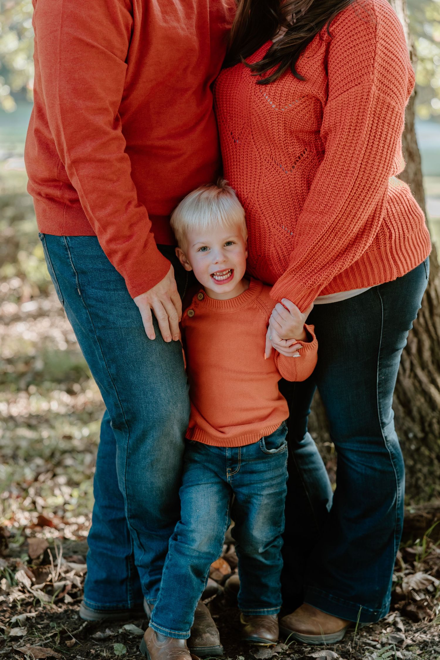 A child in an orange sweater and jeans stands close to someone wearing a coral knit sweater in a wooded setting.