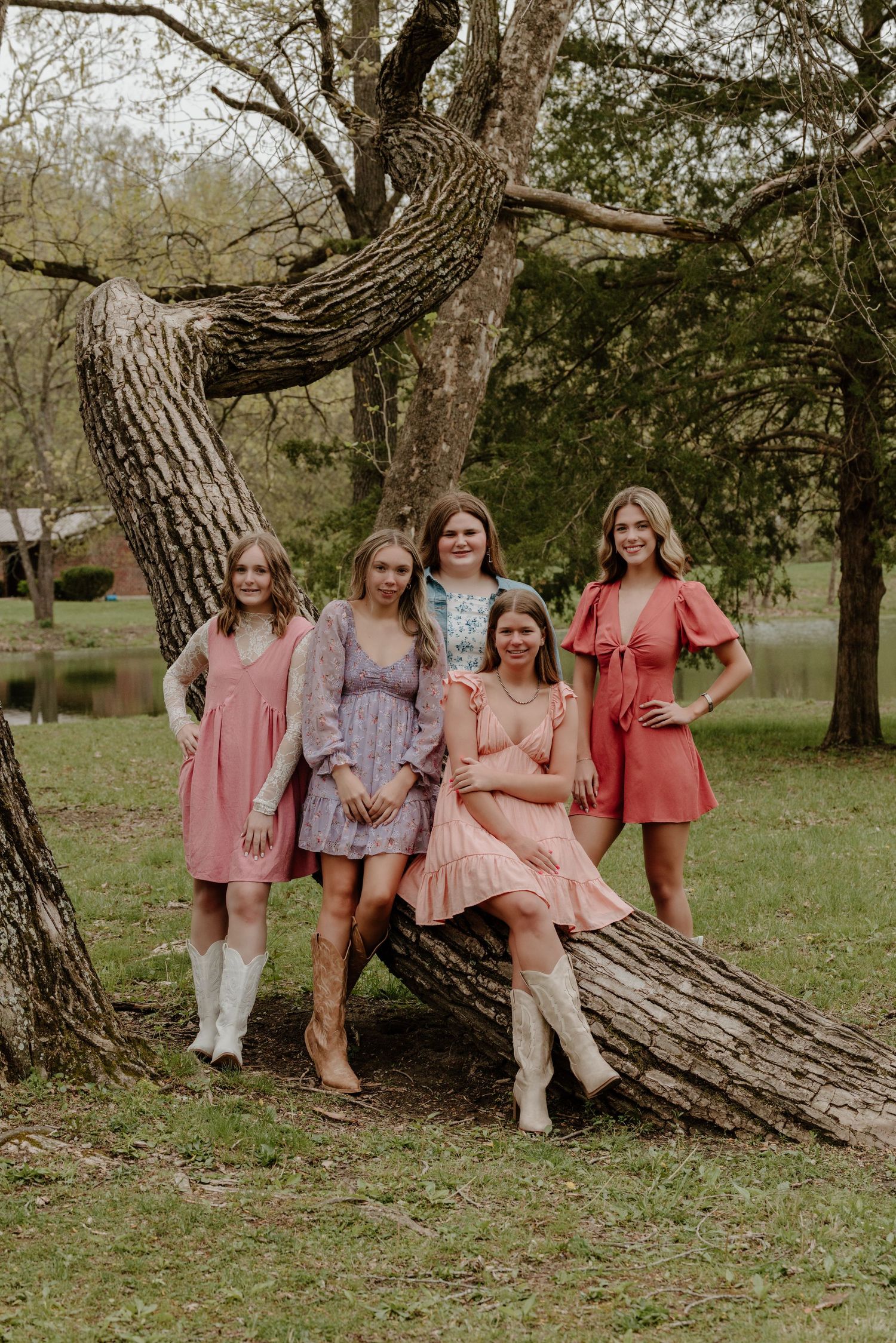 A group in coordinating pink and coral dresses pose together on a twisted tree trunk in an outdoor setting.