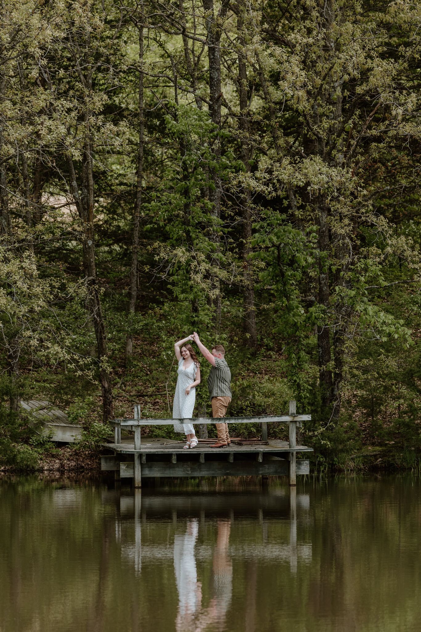 Couple dancing on a wooden dock over a calm lake surrounded by lush green forest trees.