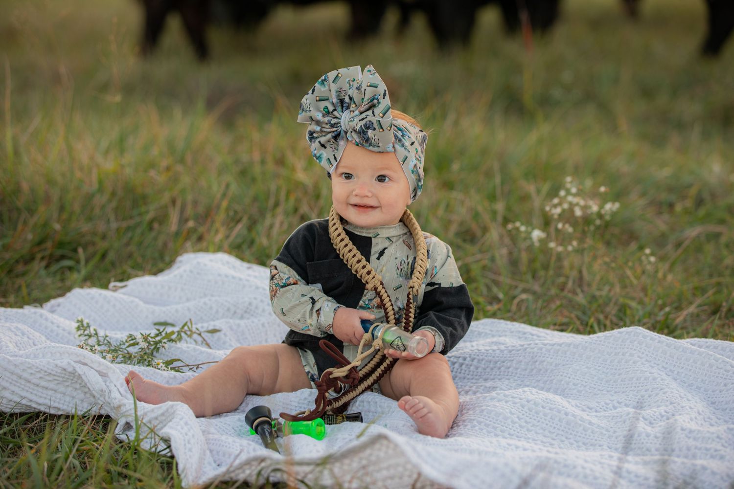 Baby in headwrap sitting on blanket outdoors with cattle in background.