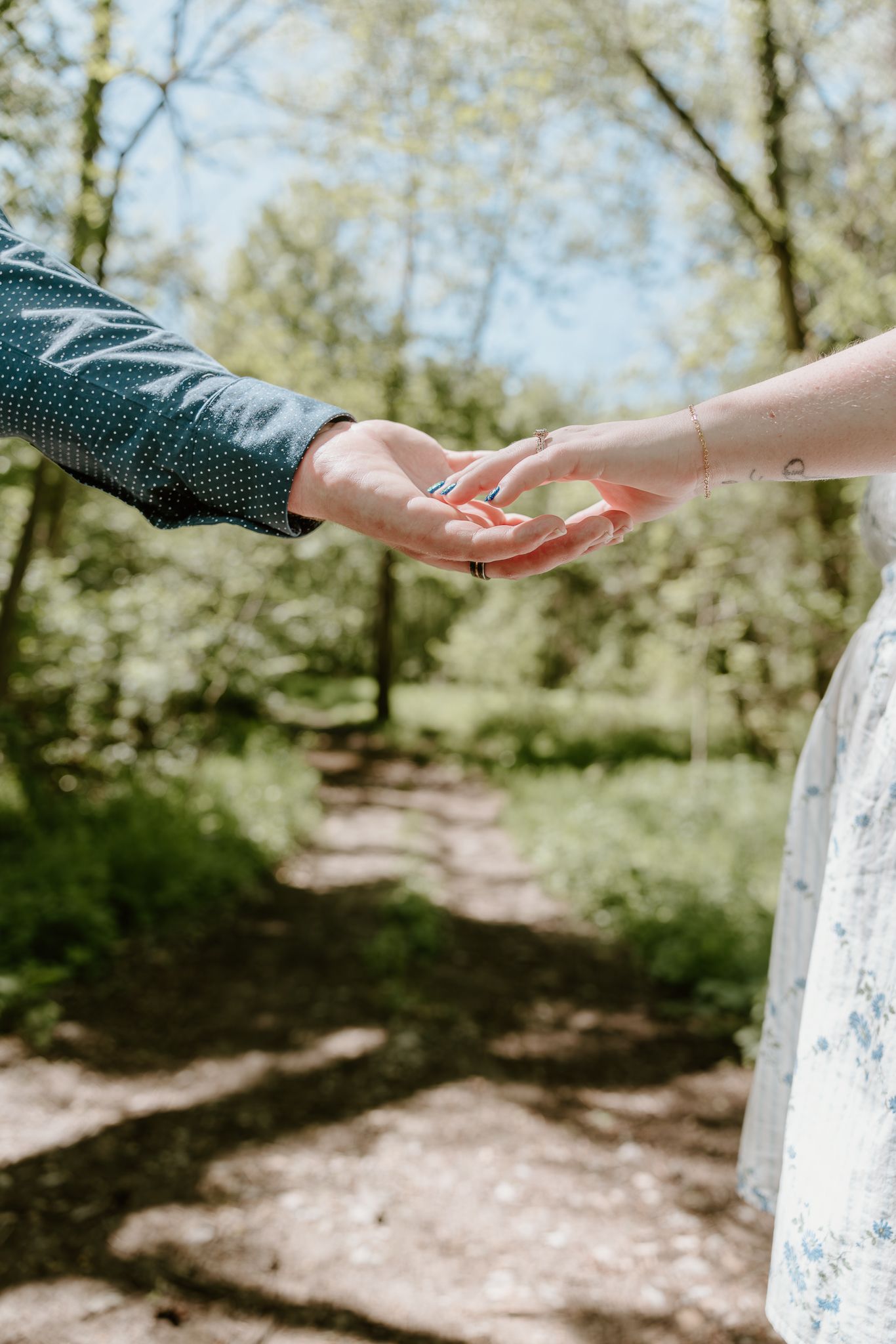 A couple gently holding hands on a sunlit forest path, surrounded by lush green trees in spring.