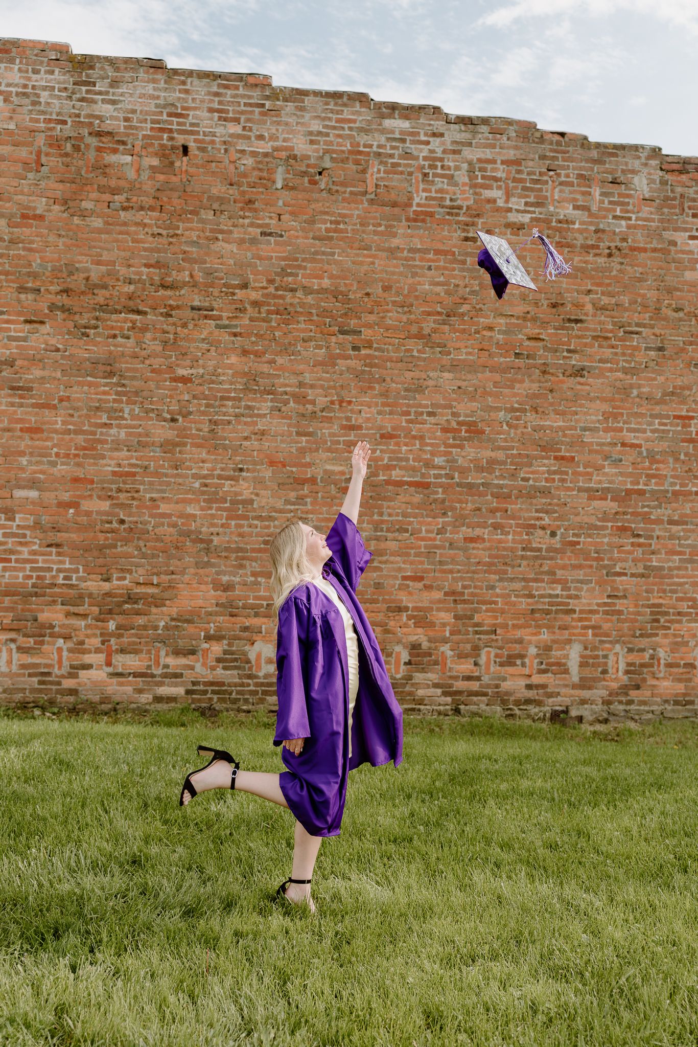 A graduate in purple cap and gown tosses her mortarboard into the air on green grass beside a brick wall.