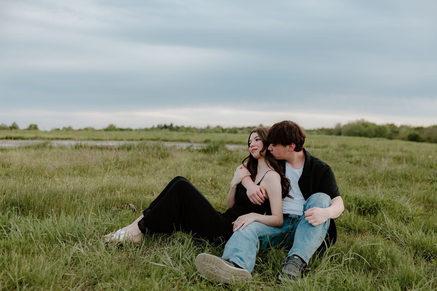 A young couple sits together on a lush green field, sharing a tender moment under a cloudy sky.