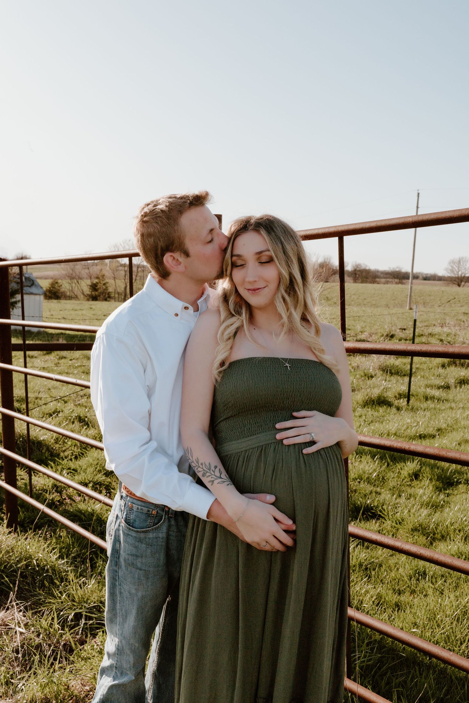 Expecting couple shares intimate moment by rustic fence in sunlit grassy field with woman wearing olive green dress.