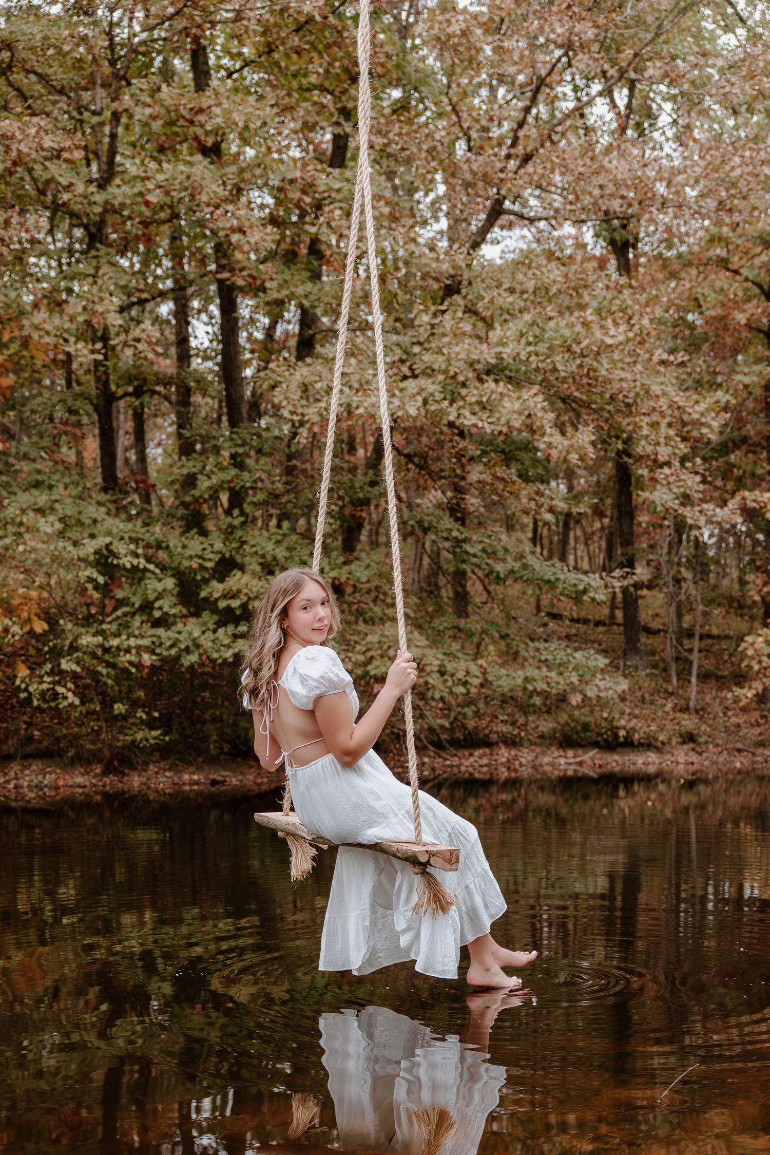 A person in a white dress sits on a rope swing over a tranquil pond surrounded by autumn trees in golden light.