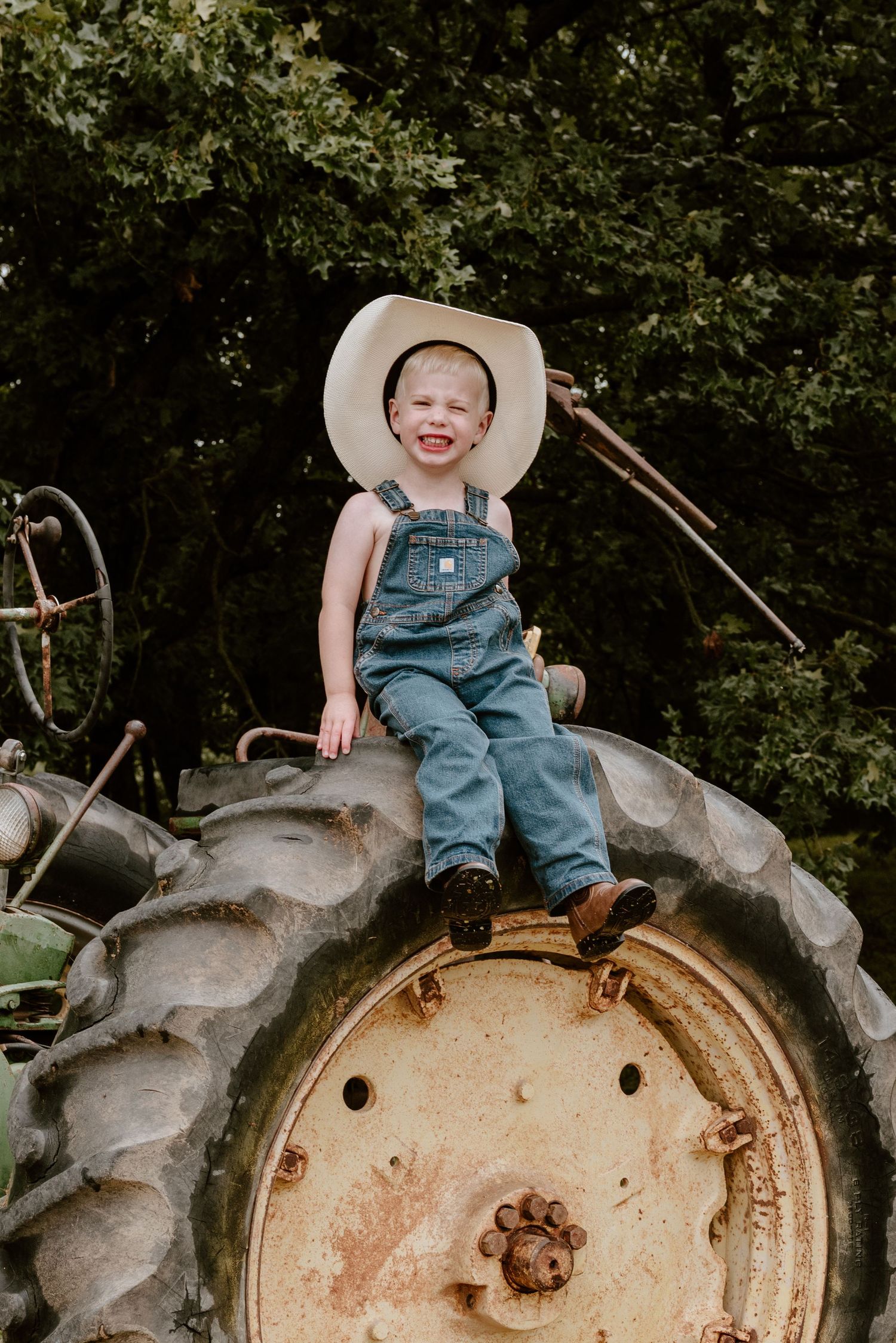 Small child in overalls and sunhat sitting on top of rustic farm tractor tire in outdoor garden setting.