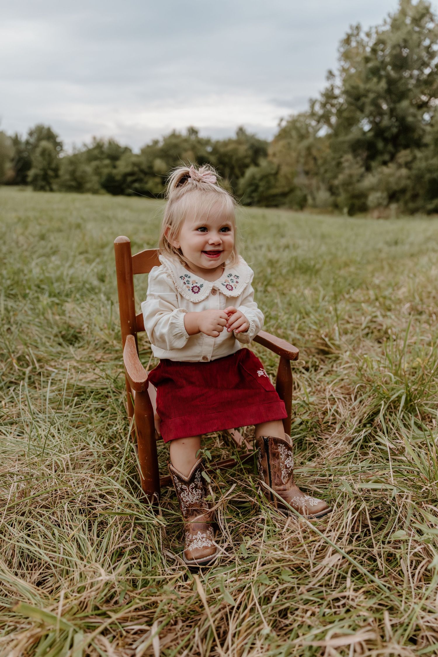 A small child sits in a wooden chair outdoors in a grassy field wearing a red dress and white collar.