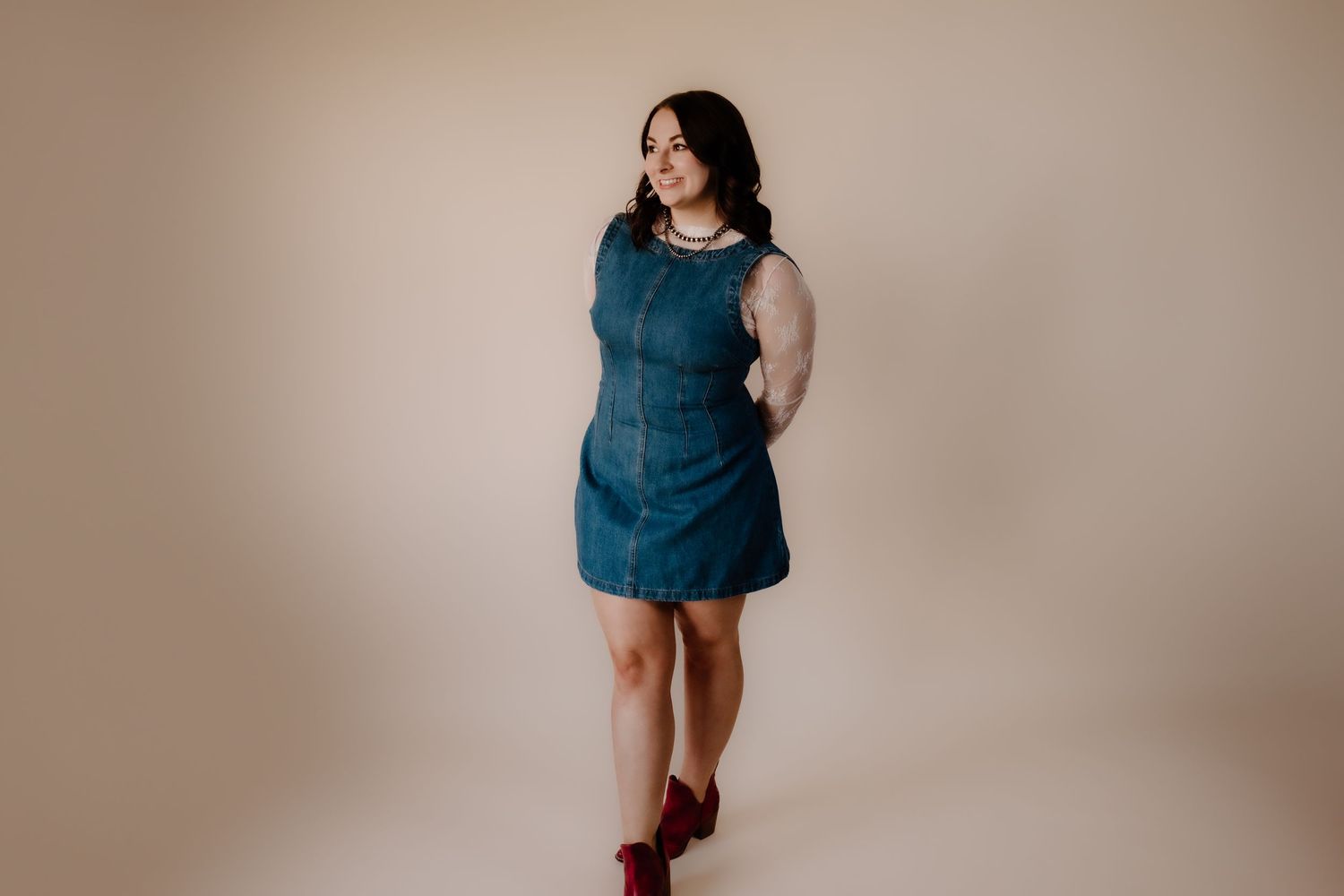 A person in a teal sleeveless dress poses against a beige background while wearing burgundy boots.