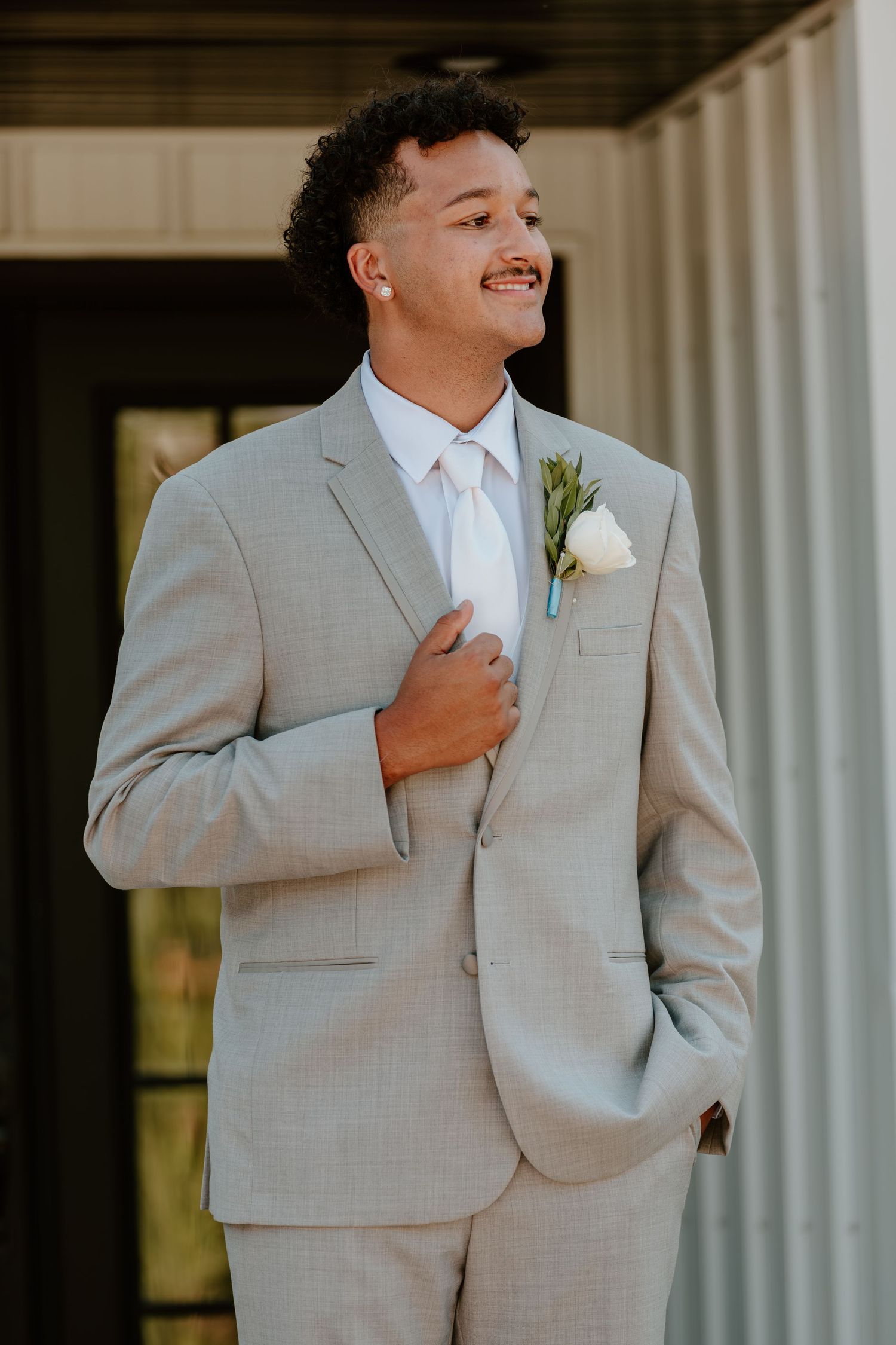 Handsome groomsman in light gray suit with white rose boutonniere smiling outdoors at wedding venue.