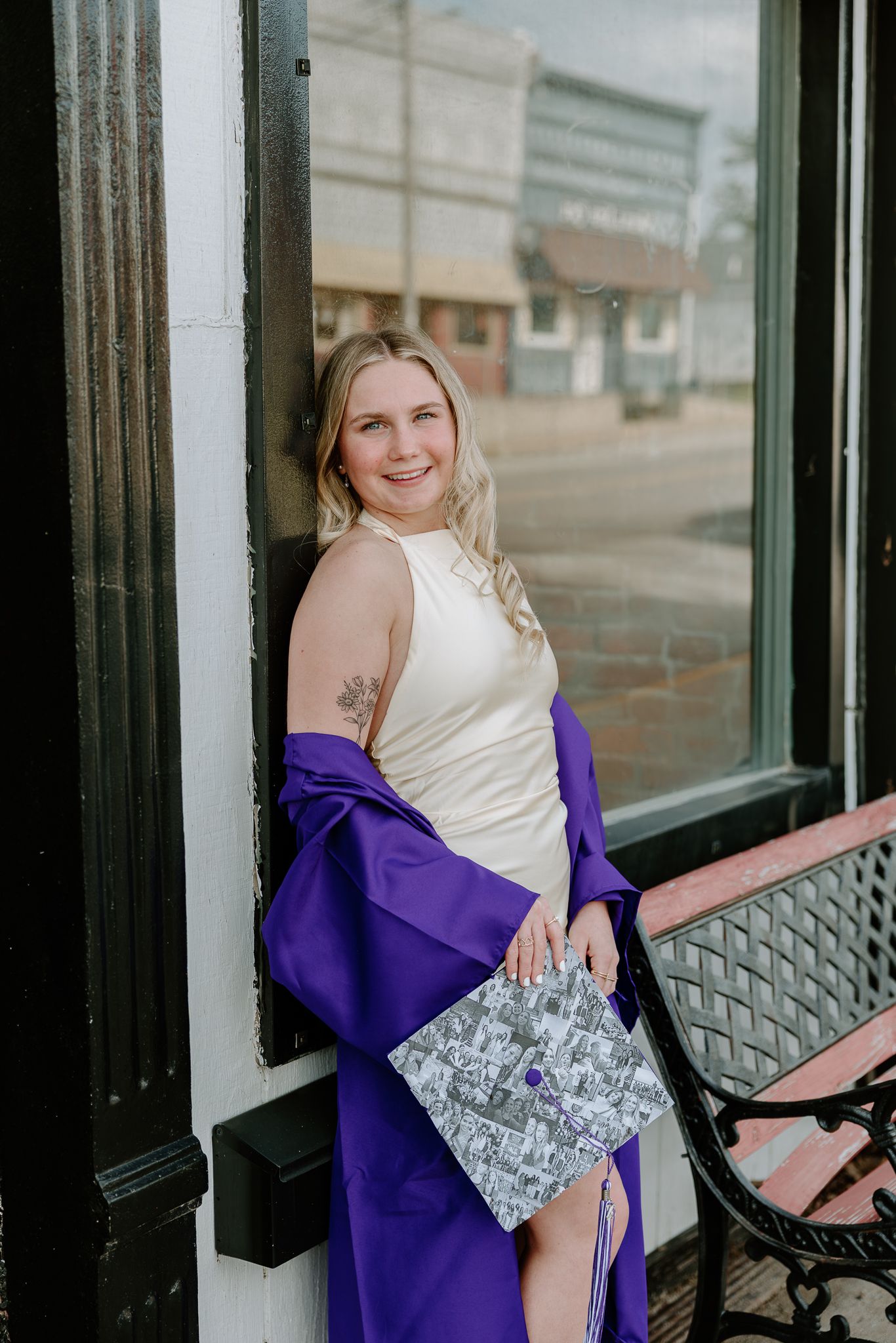 Smiling blonde woman in cream dress and purple graduation gown holding decorated cap outdoors.