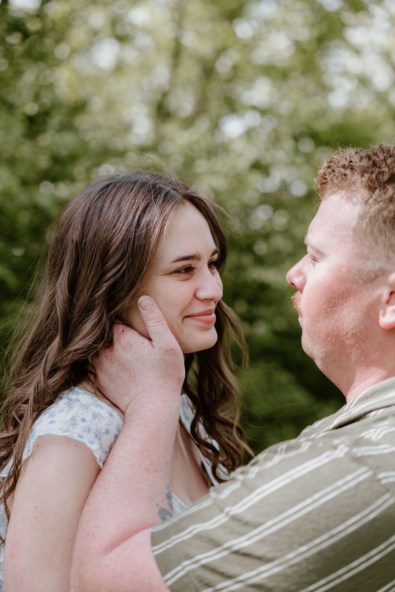 Romantic couple sharing a tender moment outdoors, man gently touching womans face near green trees.