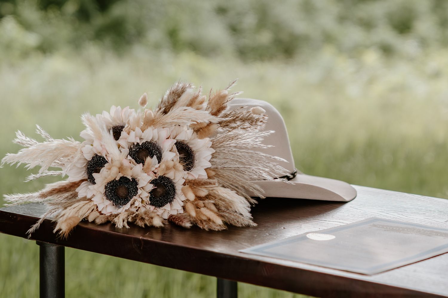A boho bouquet of dried pampas grass and sunflowers resting on a brown hat on a wooden table outdoors.
