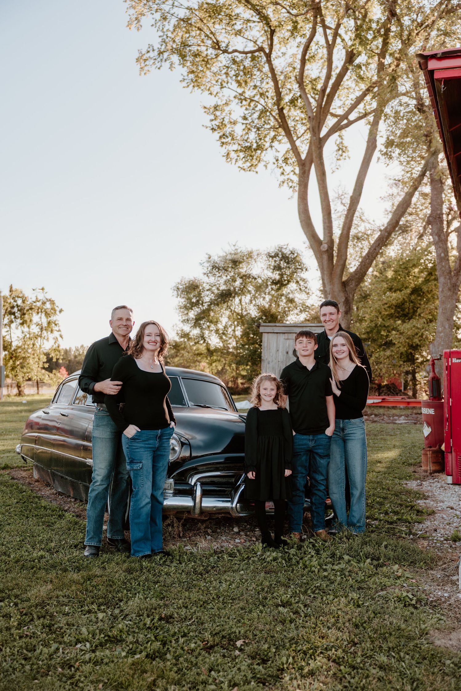 A family poses together in front of a vintage blue car on grass with trees and a red gas pump in the background.