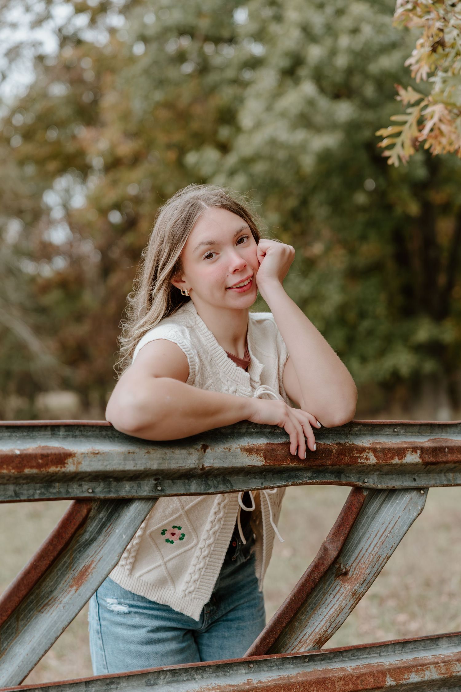 A person in jeans leans on a rustic wooden railing outdoors with autumn foliage in the background.