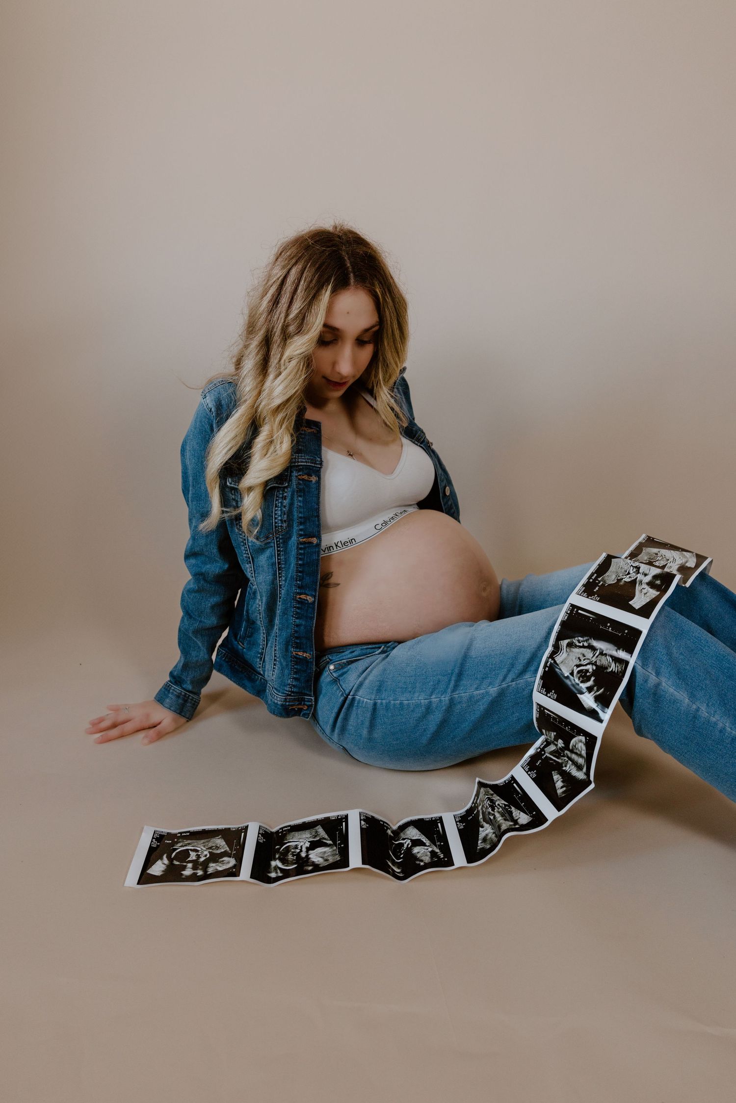 Maternity photo featuring ultrasound images arranged in a line while wearing denim jacket and jeans on beige backdrop.