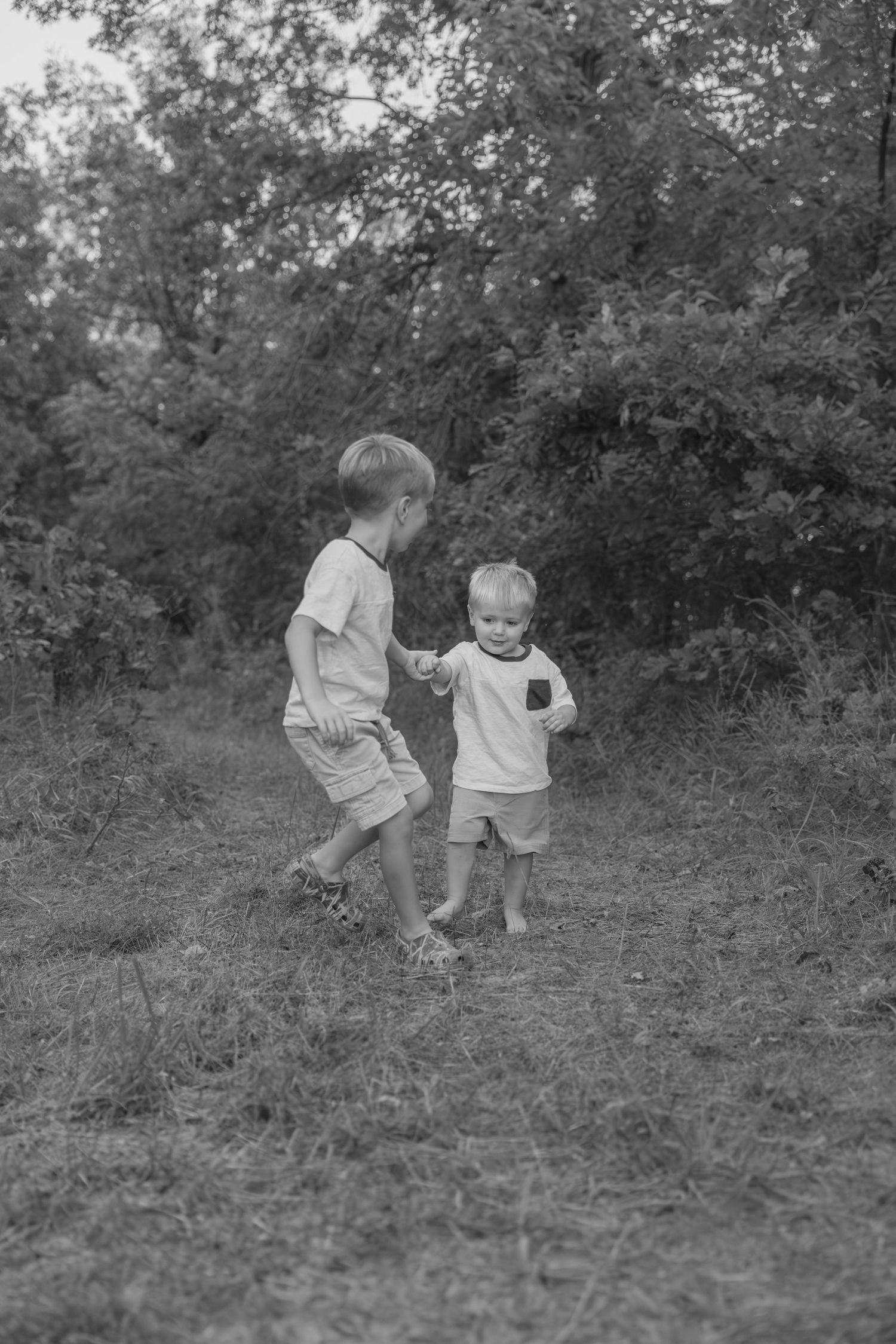 Two children in light colored clothing explore together on grassy ground in a black and white photograph.