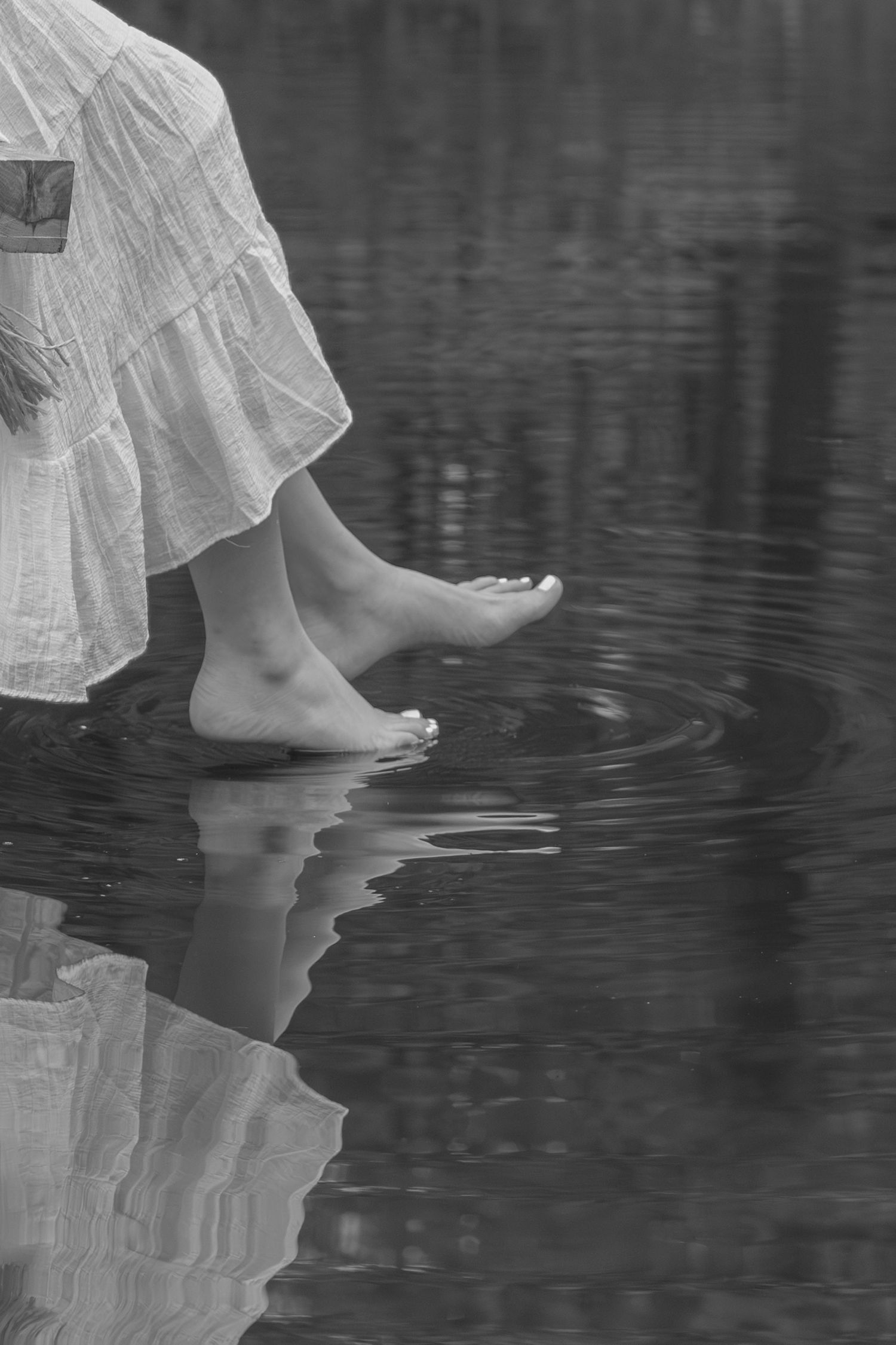 Bare feet in a white flowing dress create ripples while standing in dark reflective water.