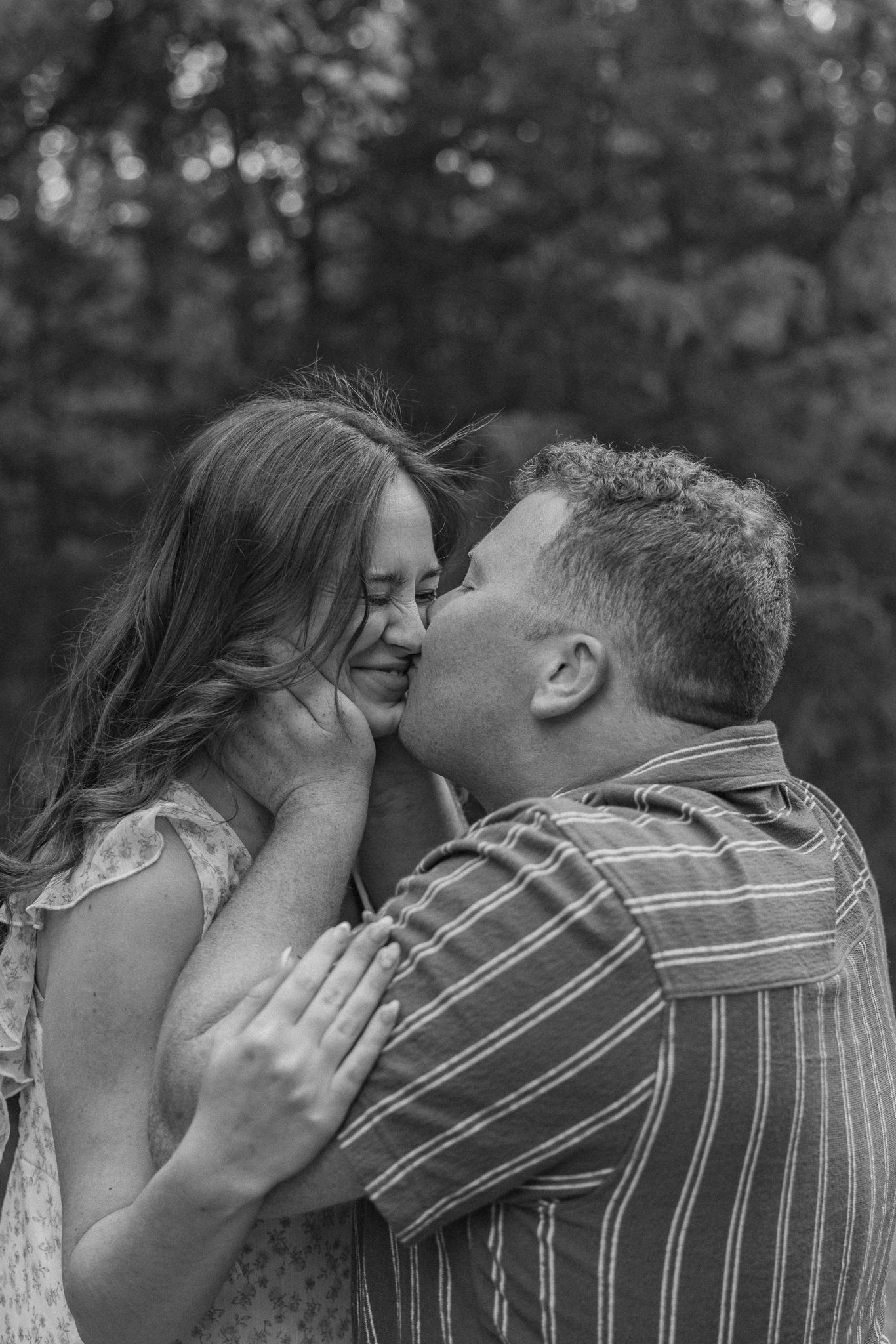 Black and white photo of a man kissing a smiling woman on the cheek outdoors in a forest setting.