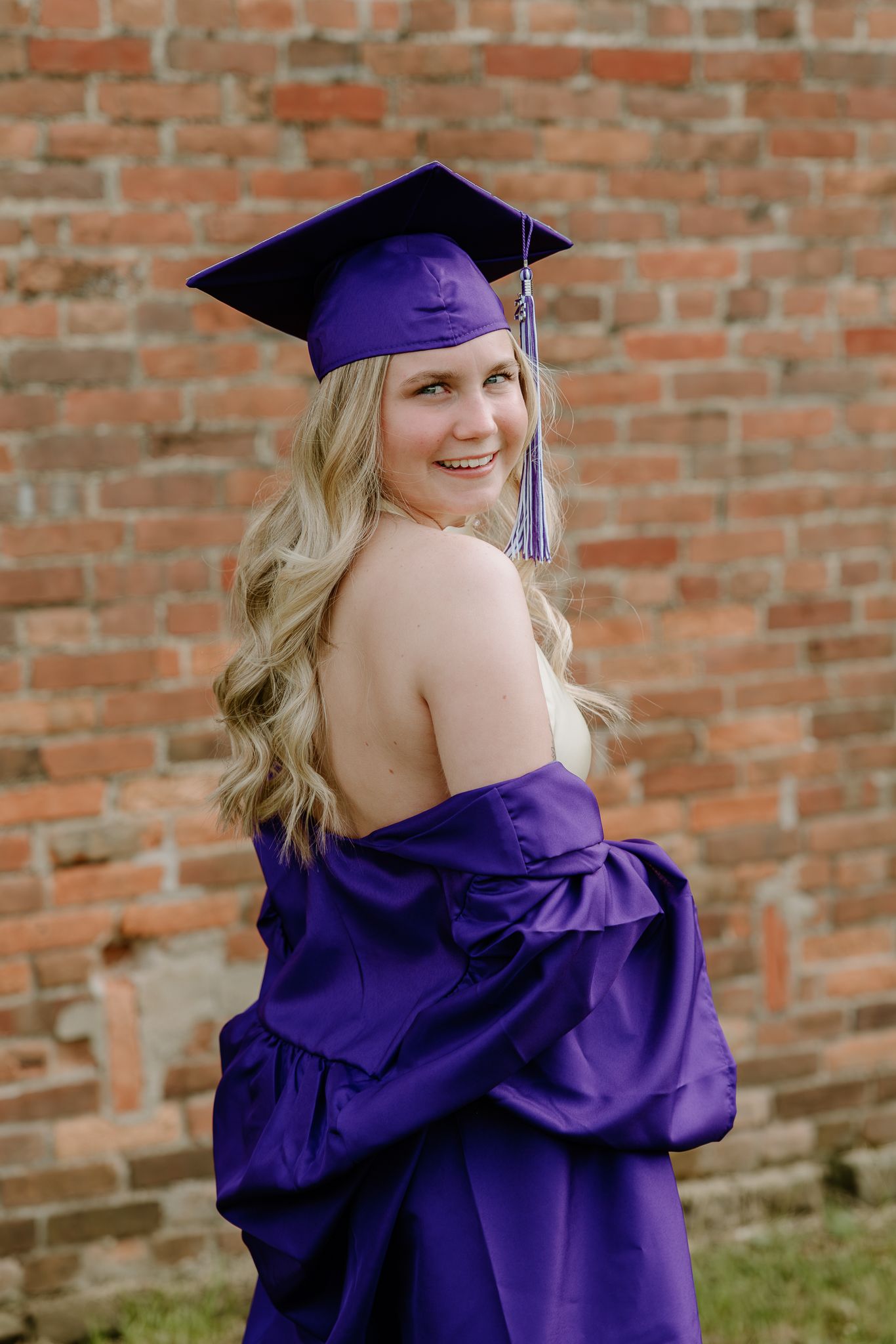 Smiling blonde graduate poses in purple cap and gown against a rustic brick wall backdrop.
